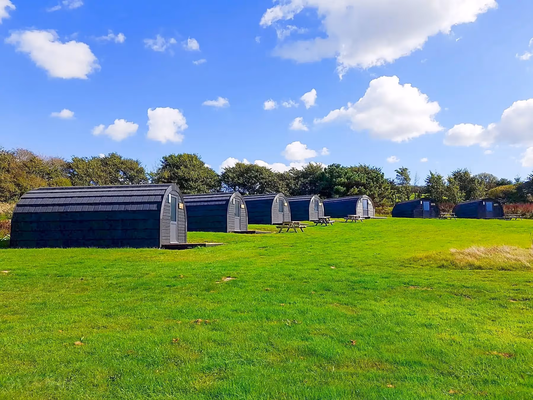 Row of modern black camping pods with picnic tables on a grassy field under a blue sky with scattered clouds.