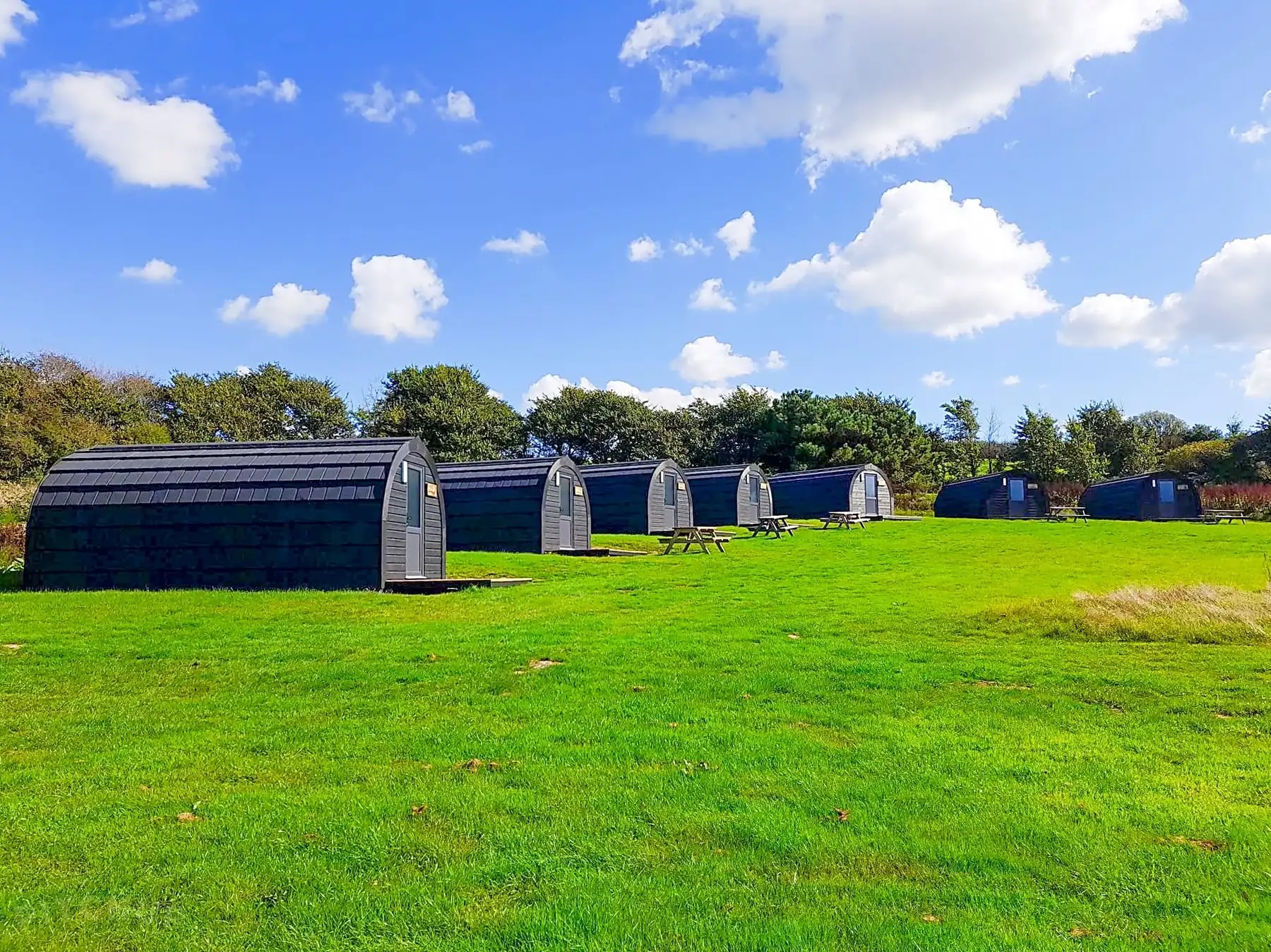 Row of modern black camping pods with picnic tables on a grassy field under a blue sky with scattered clouds.