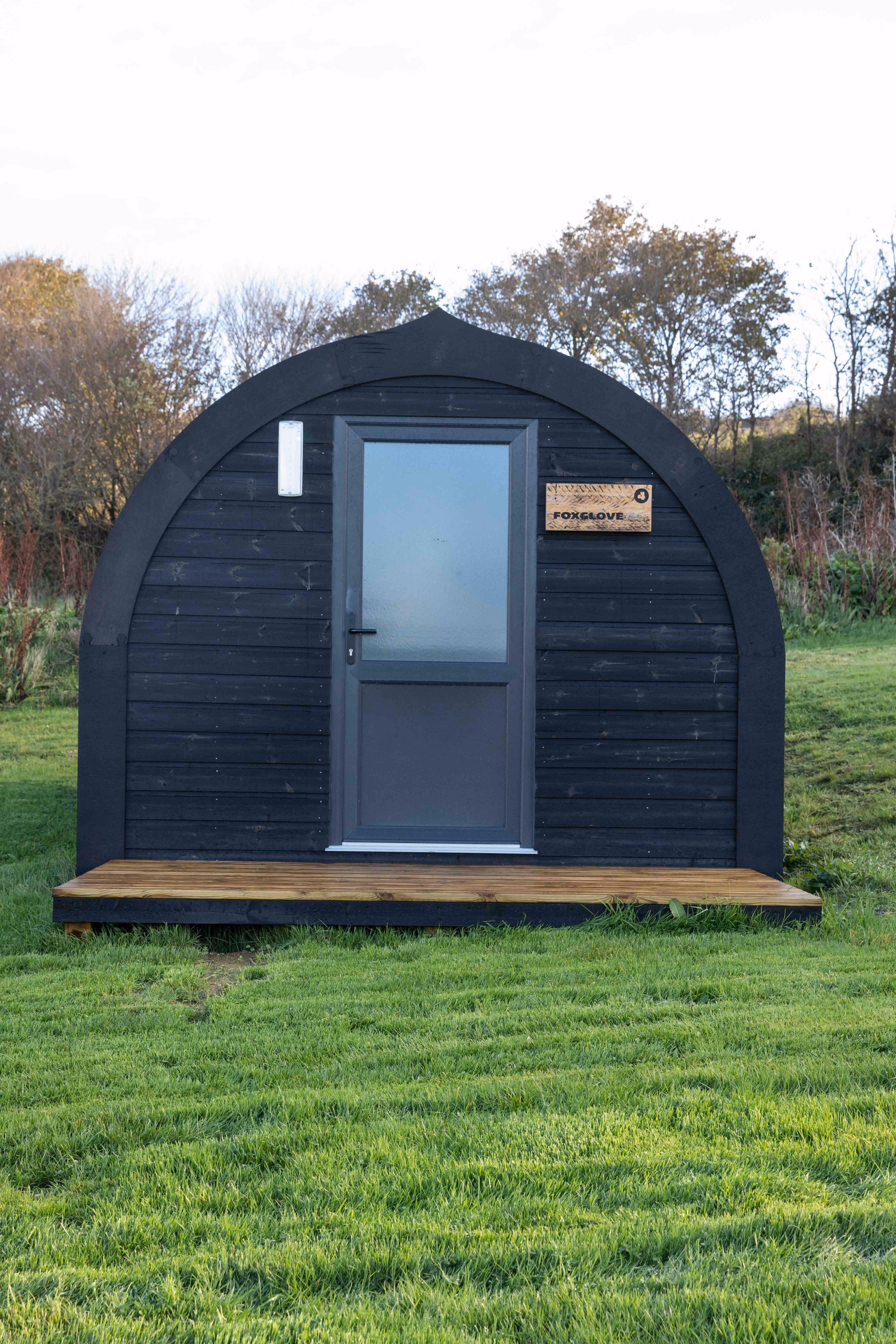 Small black wooden pod with a grey door and a wooden sign reading 'Foxglove', set on a grassy field with trees in the background.