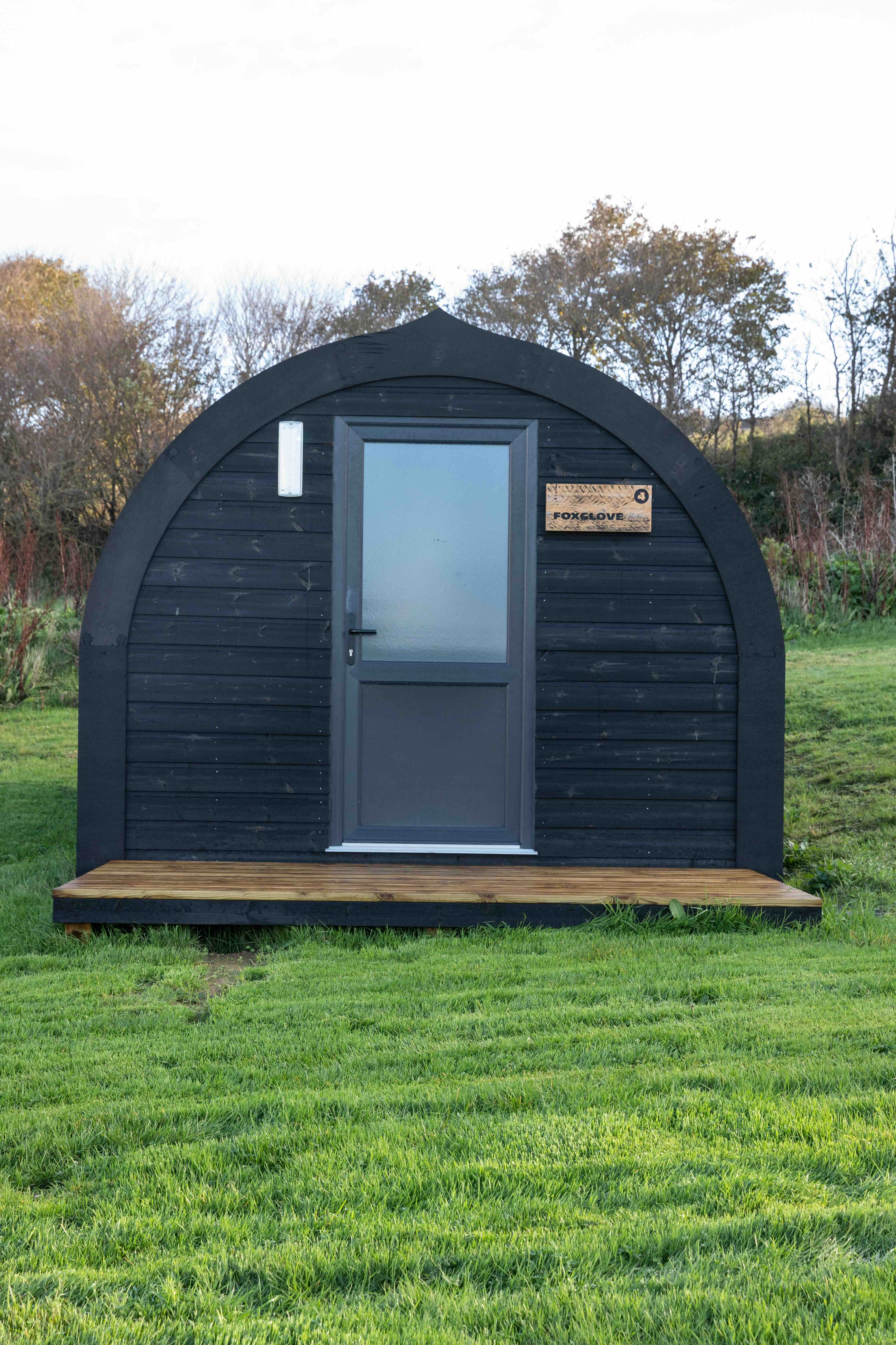 Small black wooden pod with a grey door and a wooden sign reading 'Foxglove', set on a grassy field with trees in the background.