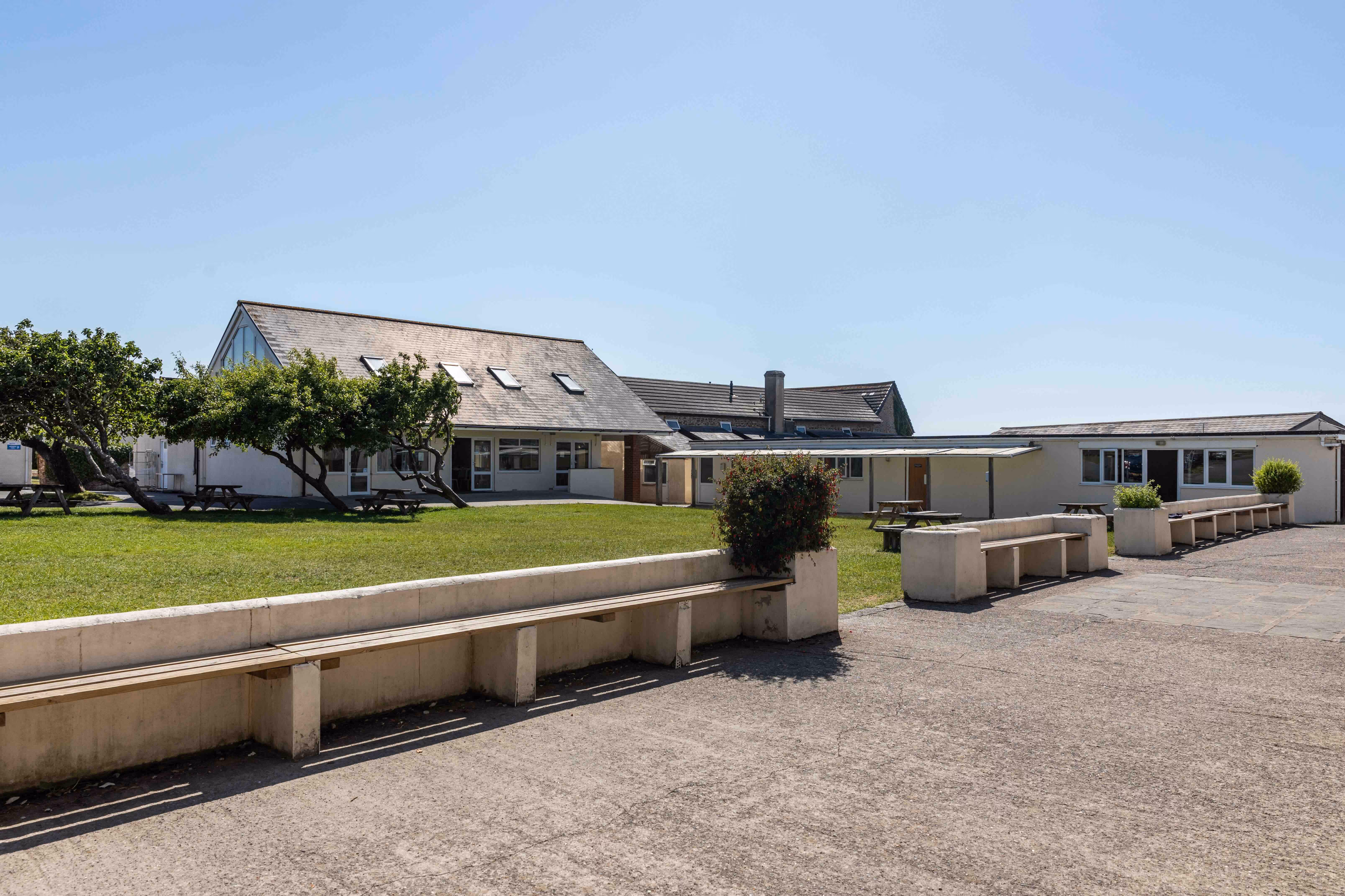 Low white buildings with pitched roofs surrounded by green grass, picnic tables, and benches under a clear blue sky.
