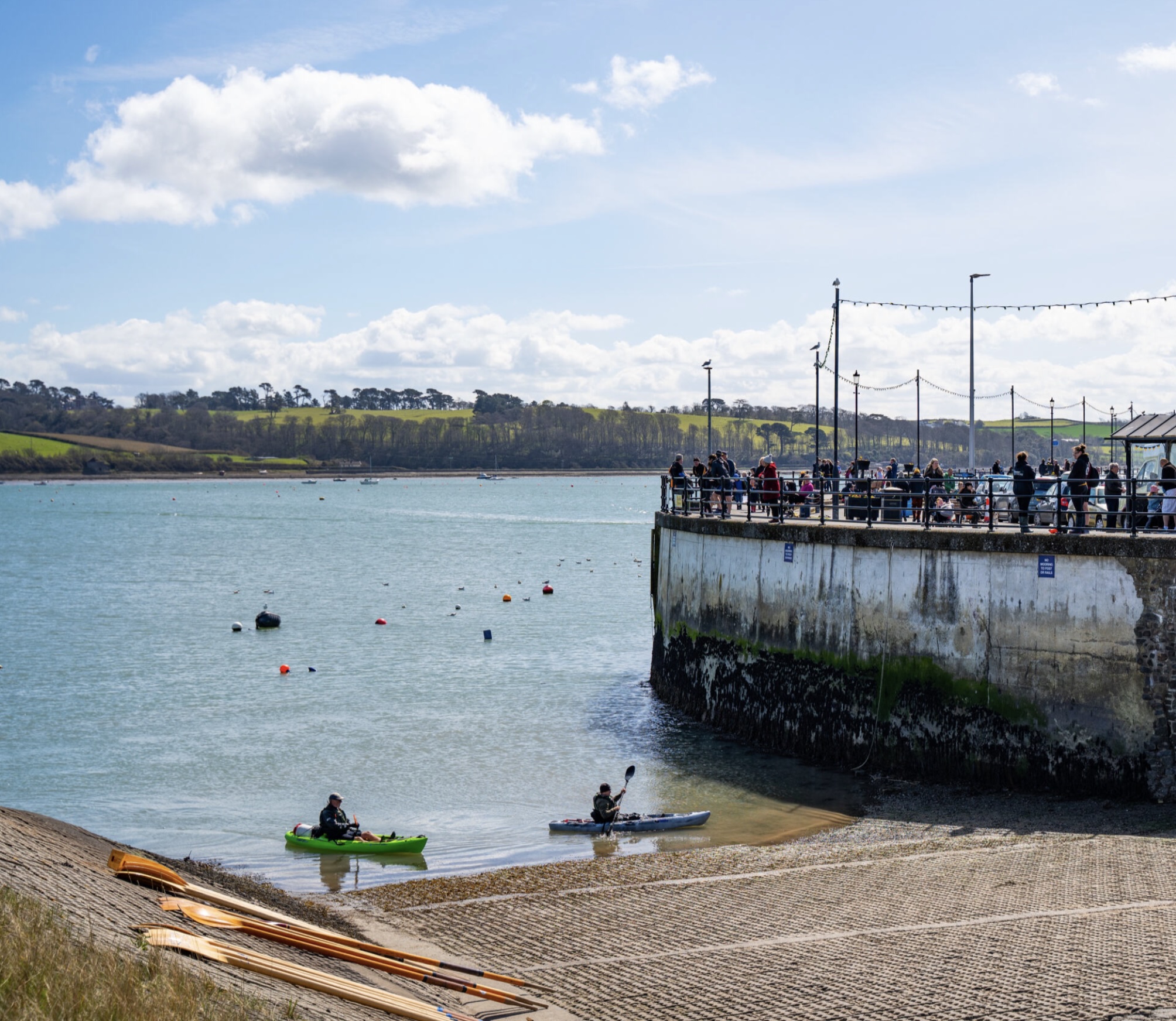 Two people kayaking near a harbour wall with a crowd of people on top and a calm sea with green hills in the background.