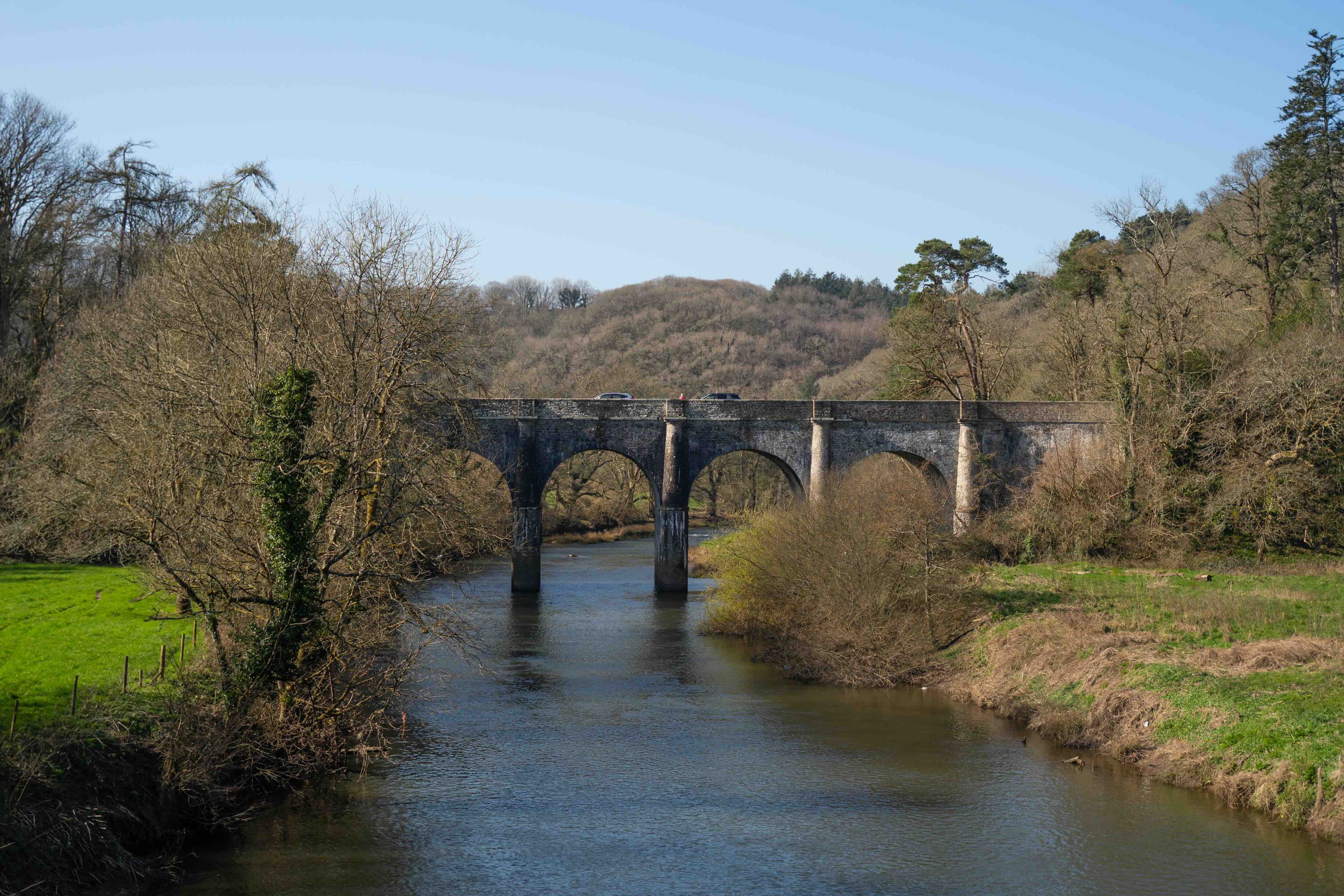 Stone arch bridge spanning a calm river with leafless trees and grassy banks under a clear blue sky.