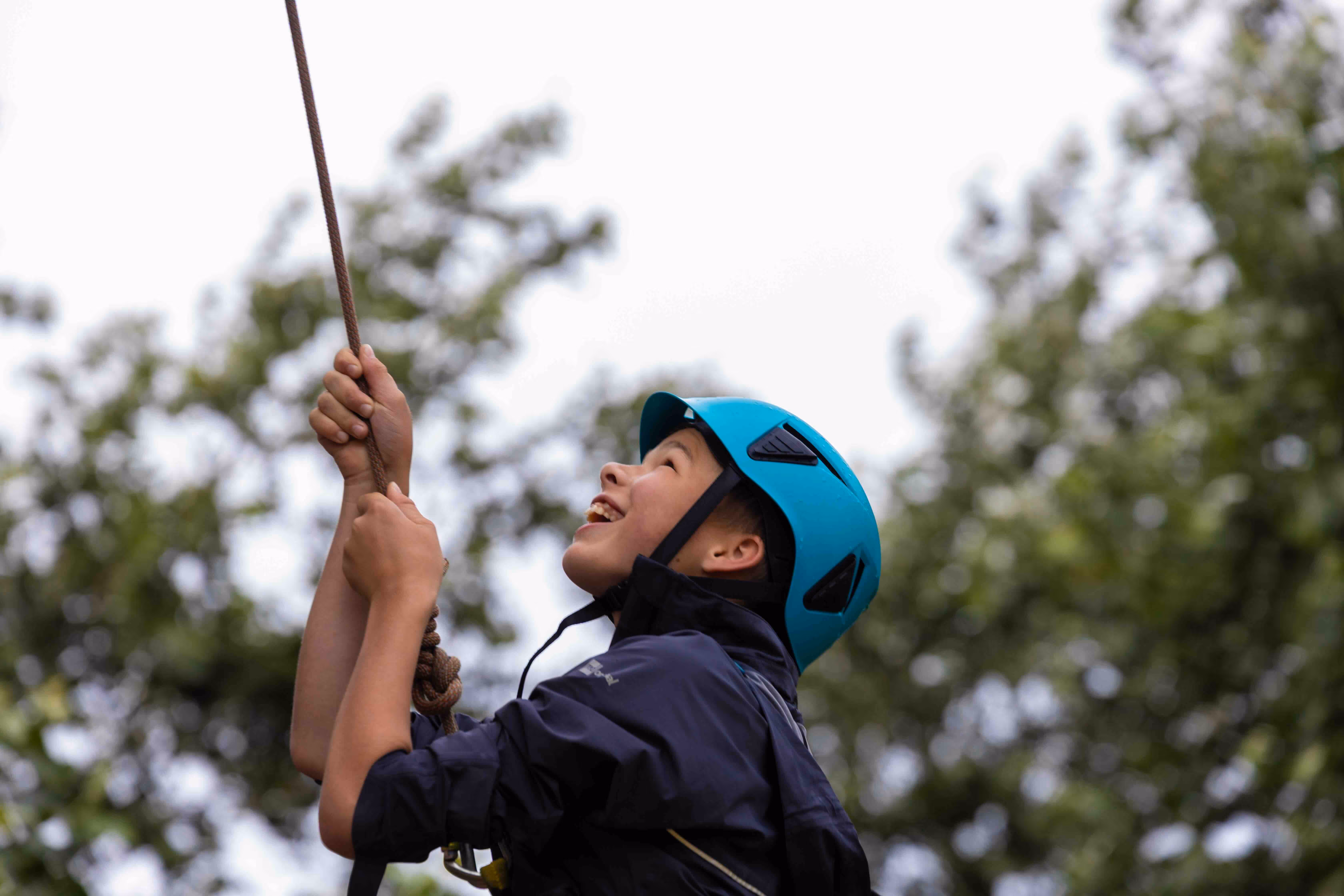 Boy wearing a blue helmet and dark jacket climbing with a rope outdoors.