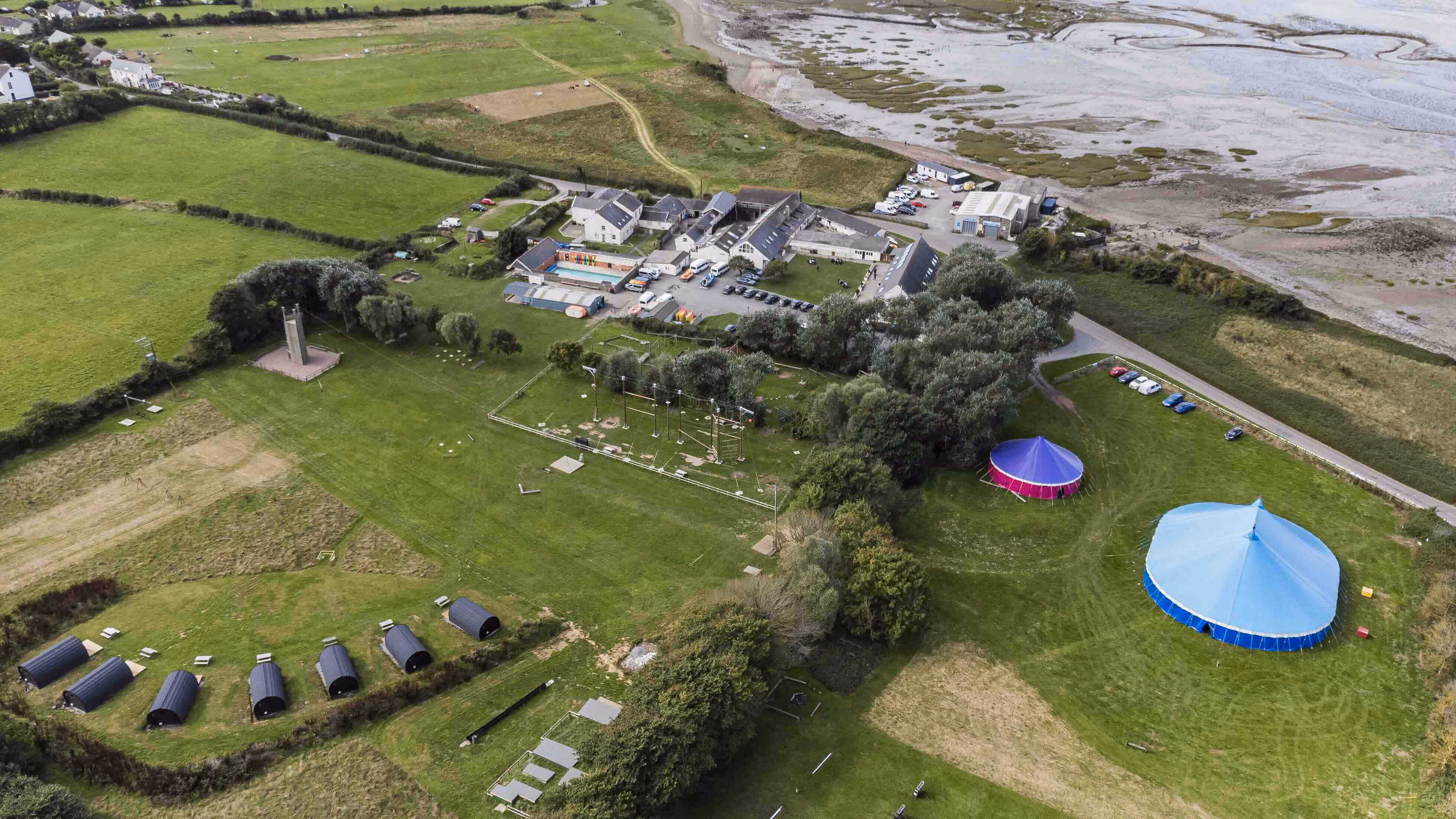 Aerial view of a coastal outdoor activity centre with tents, cabins, climbing structures, and green fields near a mudflat shore.