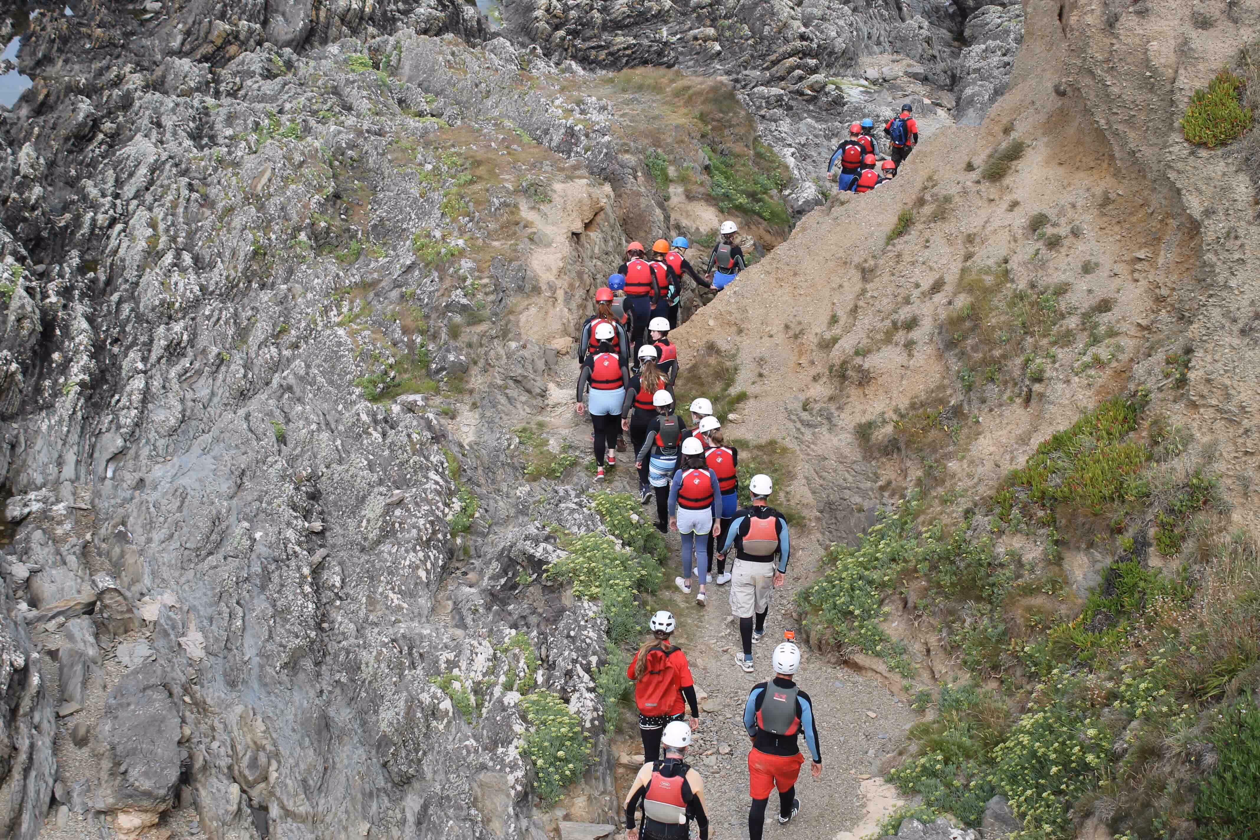 Group of people wearing helmets and wetsuits walking in a line along a narrow rocky coastal path.