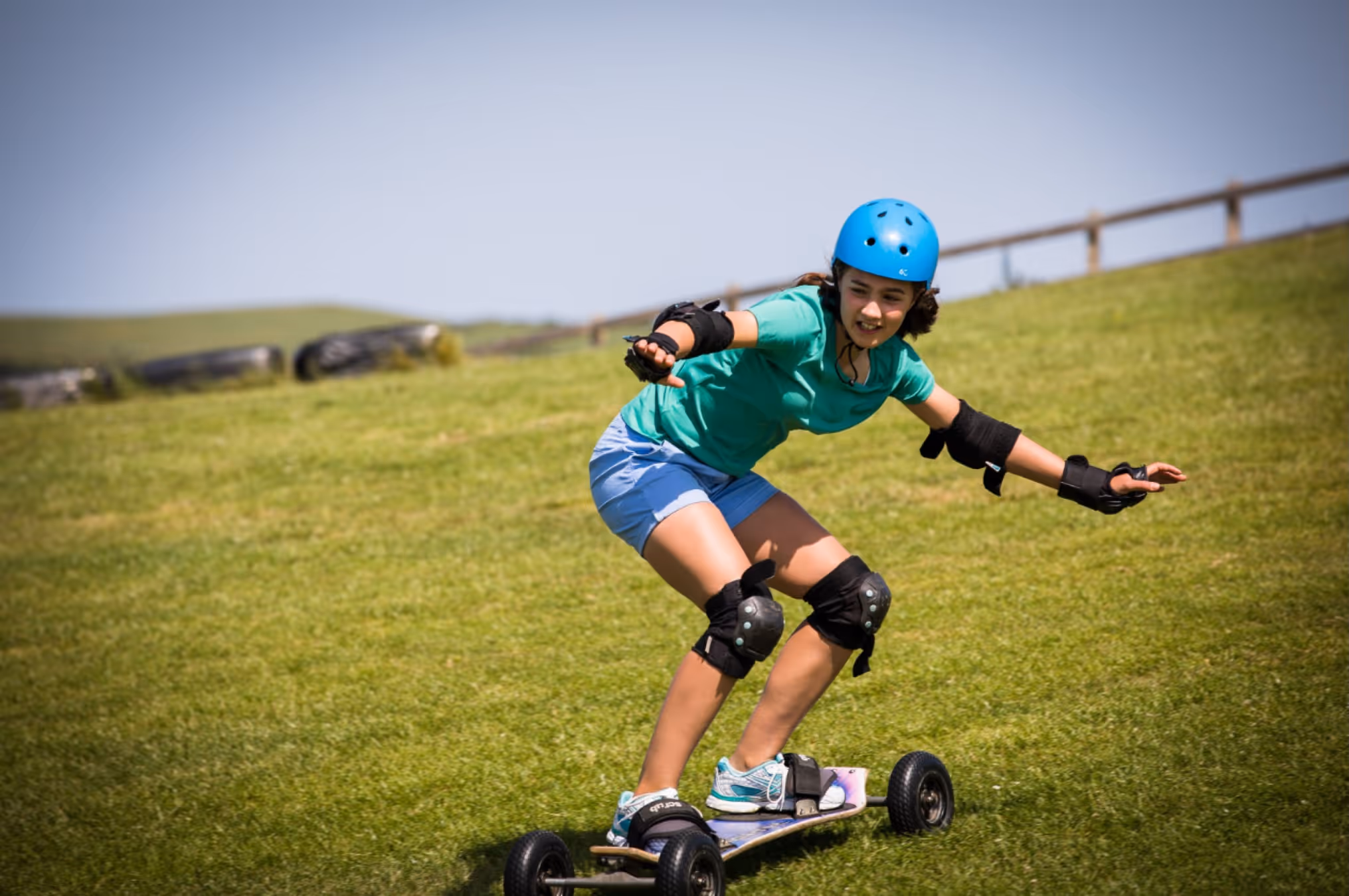 Person wearing protective gear and blue helmet riding a mountain board downhill on grass.