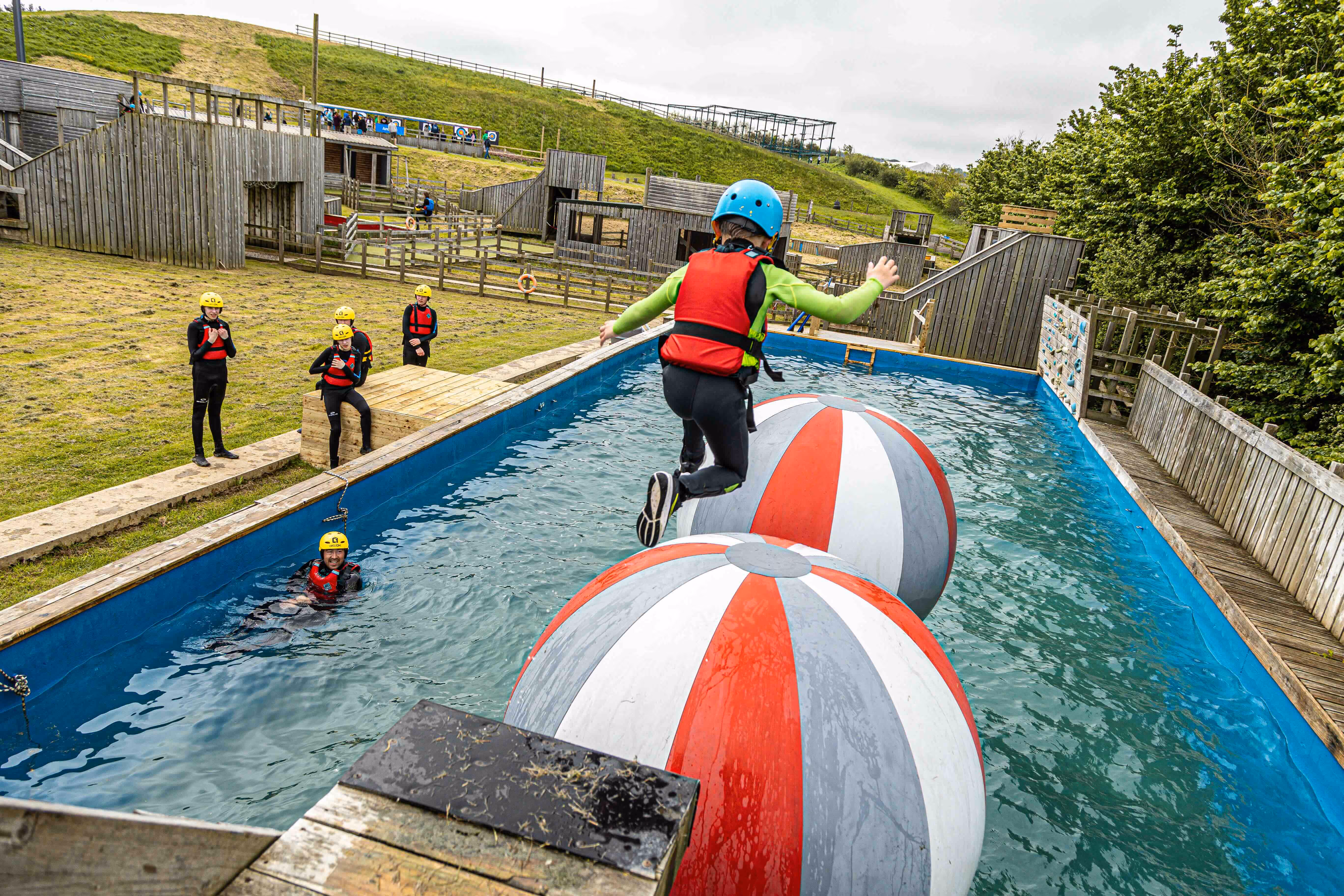 Child wearing helmet and life jacket jumping across large inflatable balls in a water obstacle course while others watch.