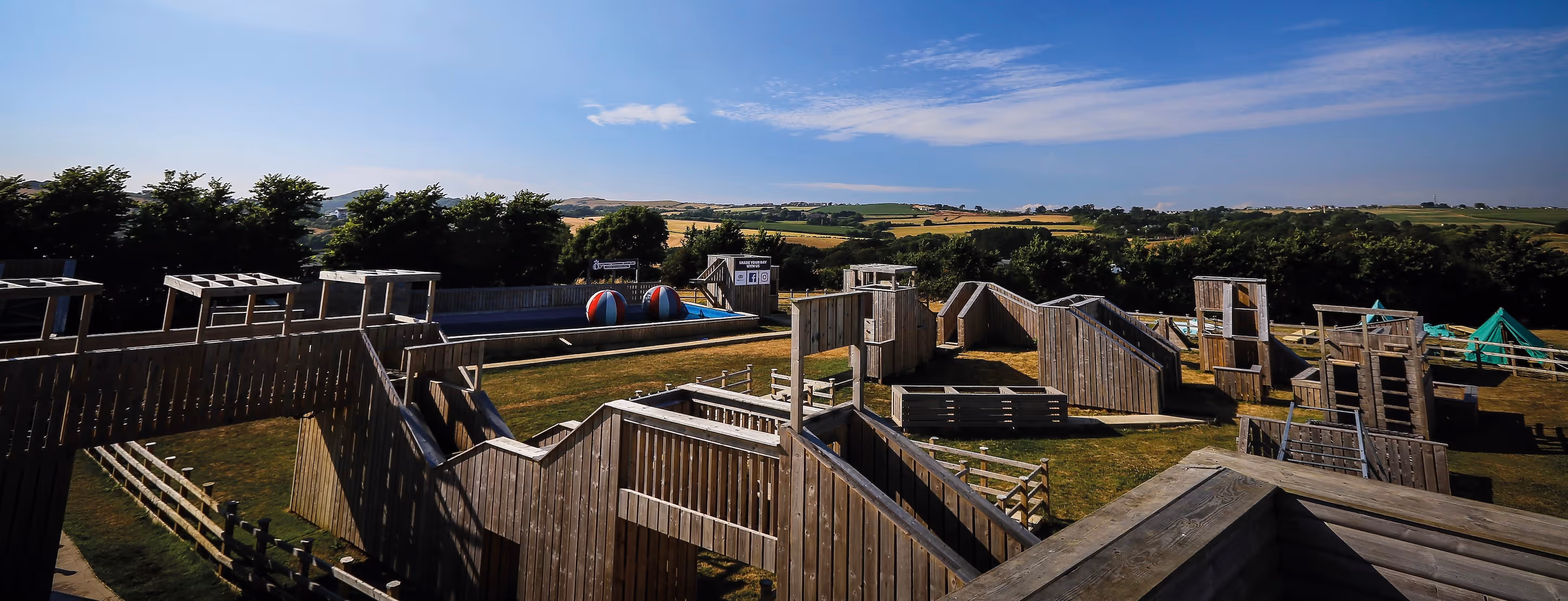Wooden outdoor adventure course with obstacles and two large inflatable balls in a pool, set in a countryside landscape under a blue sky.