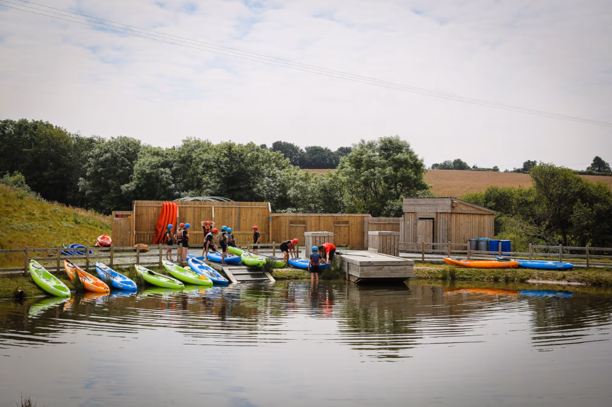 Group of people in helmets by a lakeside with colourful kayaks and wooden docks in a rural outdoor setting.