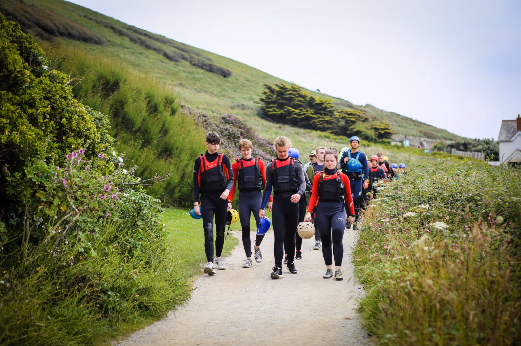 Group of people wearing wetsuits and life jackets walking on a path surrounded by green hills and bushes.