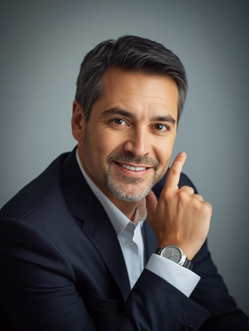 Smiling middle-aged man with gray hair and beard wearing a dark suit and watch, pointing upward with his index finger.