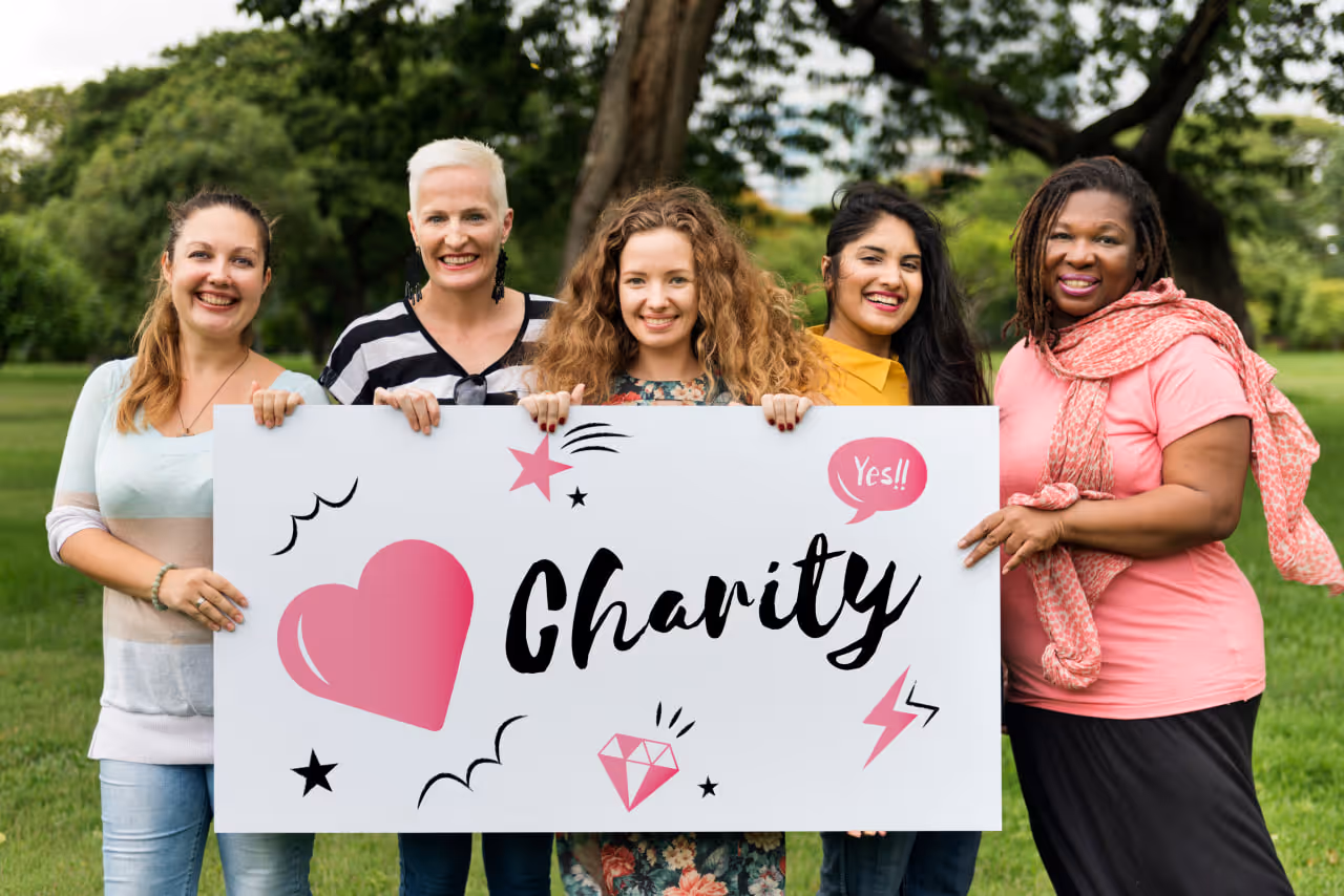 Five diverse women standing outdoors holding a sign that says 'Charity' with pink heart and diamond graphics.