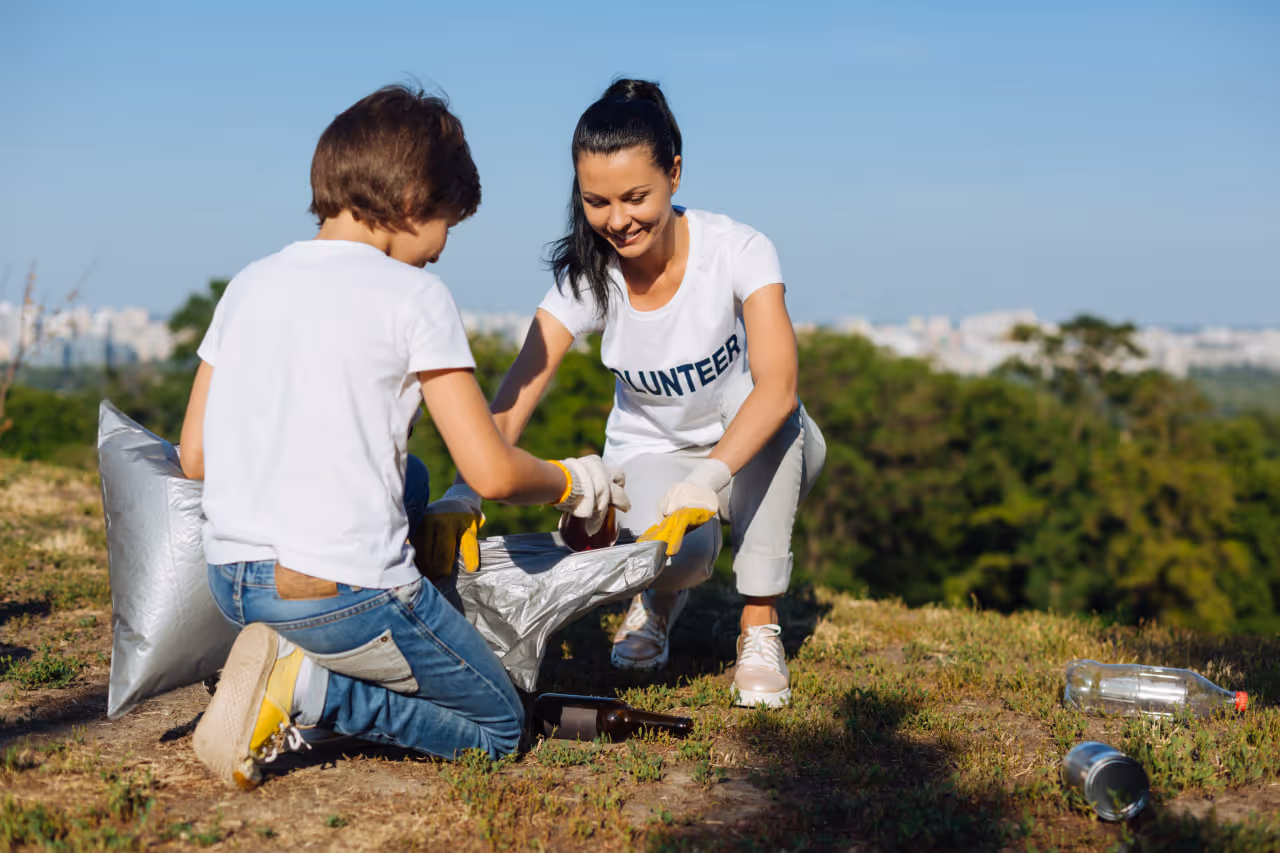 A woman in a volunteer t-shirt and a boy collecting trash outdoors on a grassy area.