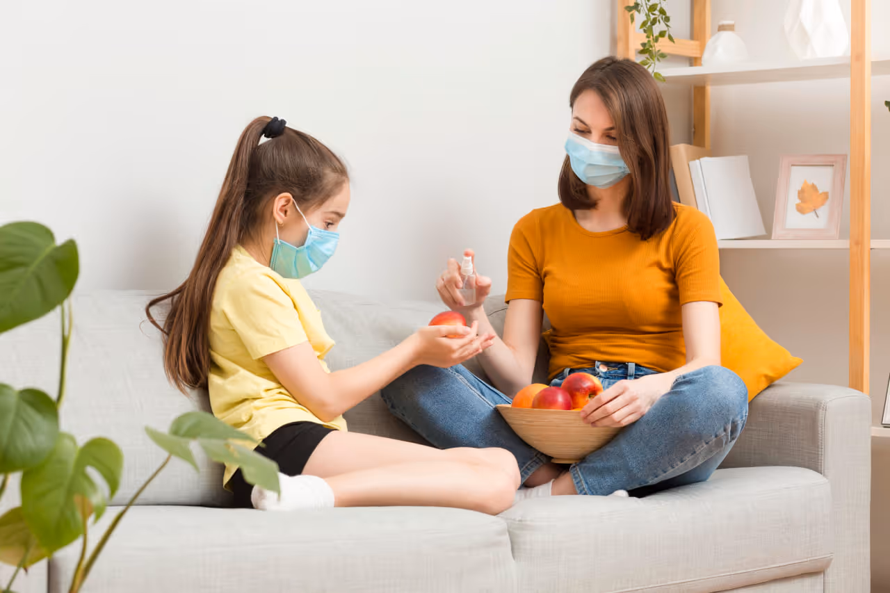 Woman in an orange shirt spraying sanitizer on a girl's hands while both wear face masks and sit on a couch holding apples.