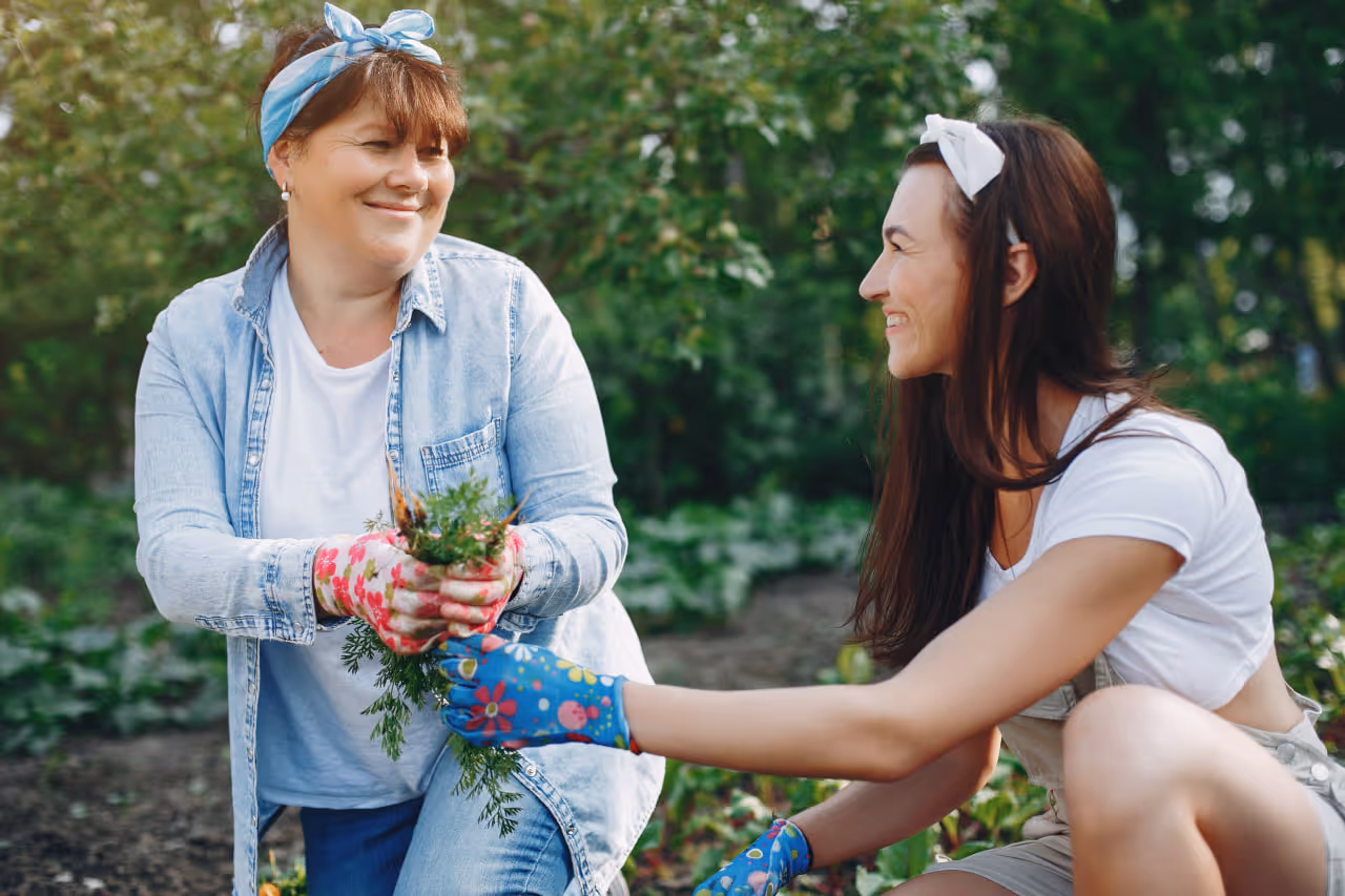 Two women wearing gardening gloves and headbands smiling at each other while holding freshly picked carrots in a garden.