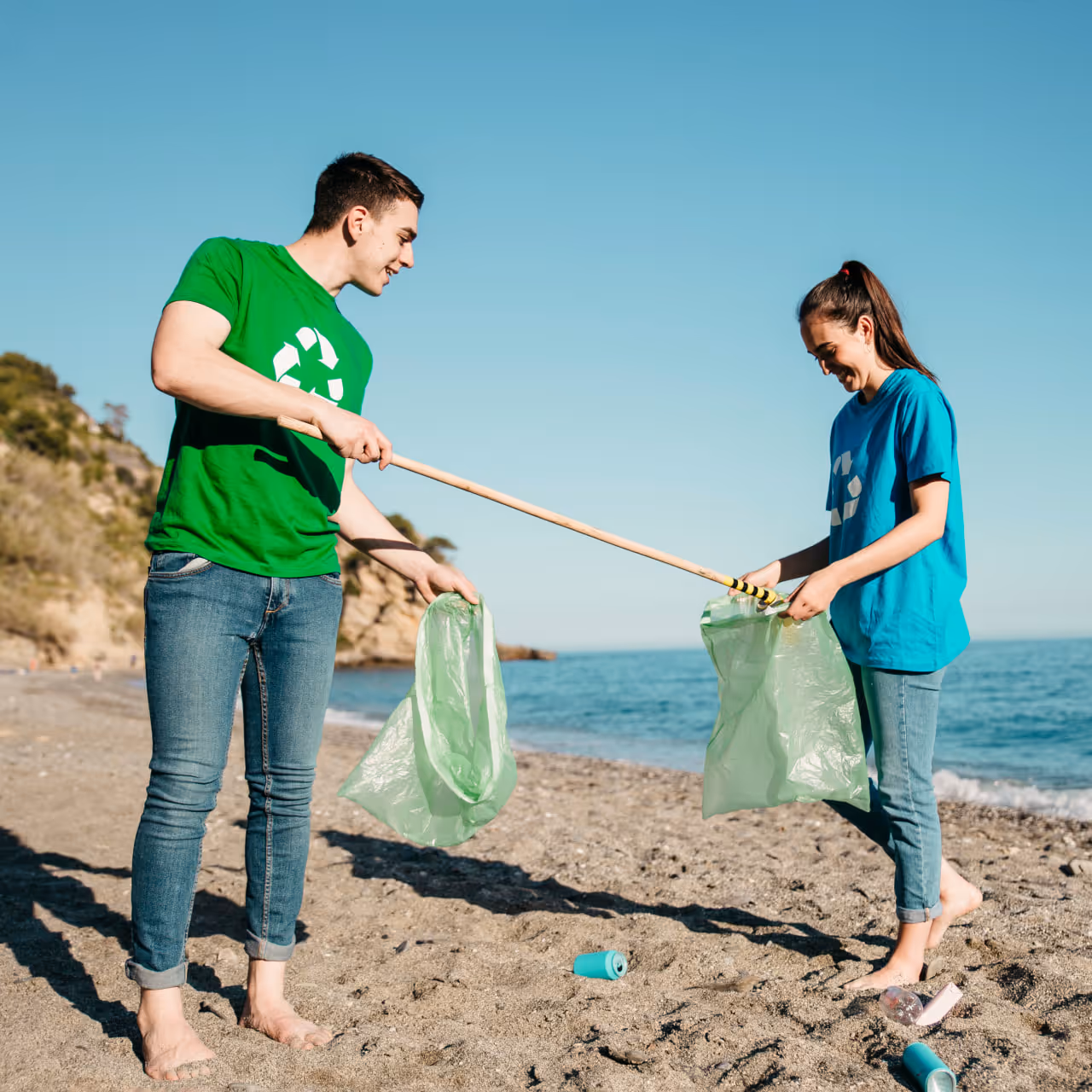 Two young people wearing recycling symbol shirts picking up trash on a beach with large green bags and a grabber tool.