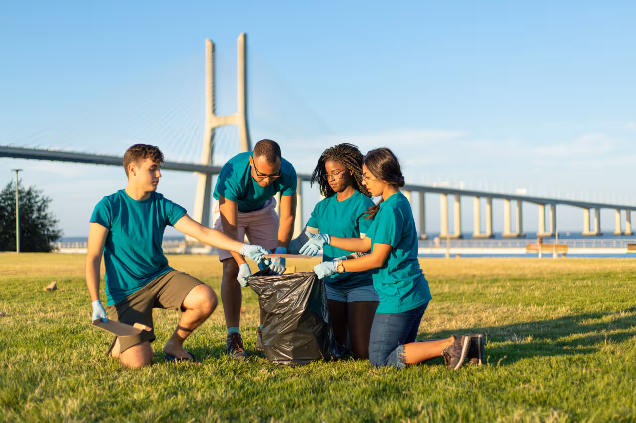 Four volunteers in teal shirts cleaning up a park near a large suspension bridge, placing trash into a black garbage bag.