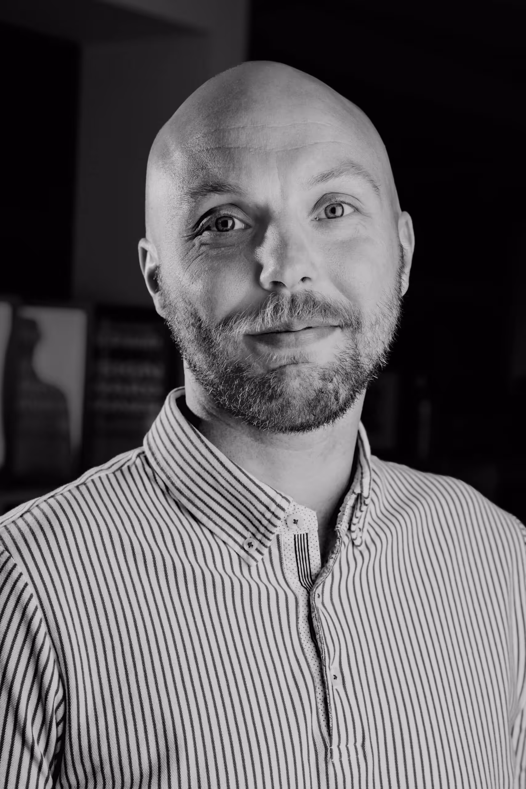 Black and white portrait of a bald man with a beard wearing a striped button-up shirt, smiling slightly.