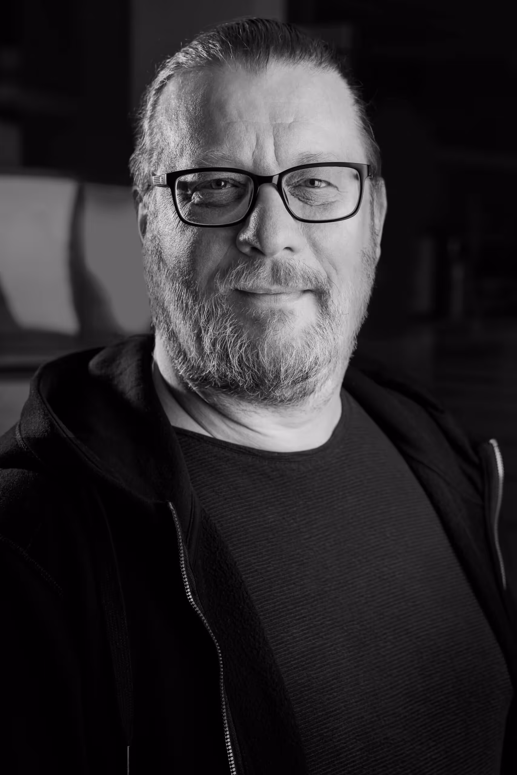 Black and white portrait of a middle-aged man with glasses, beard, and a slight smile wearing a dark shirt and jacket.