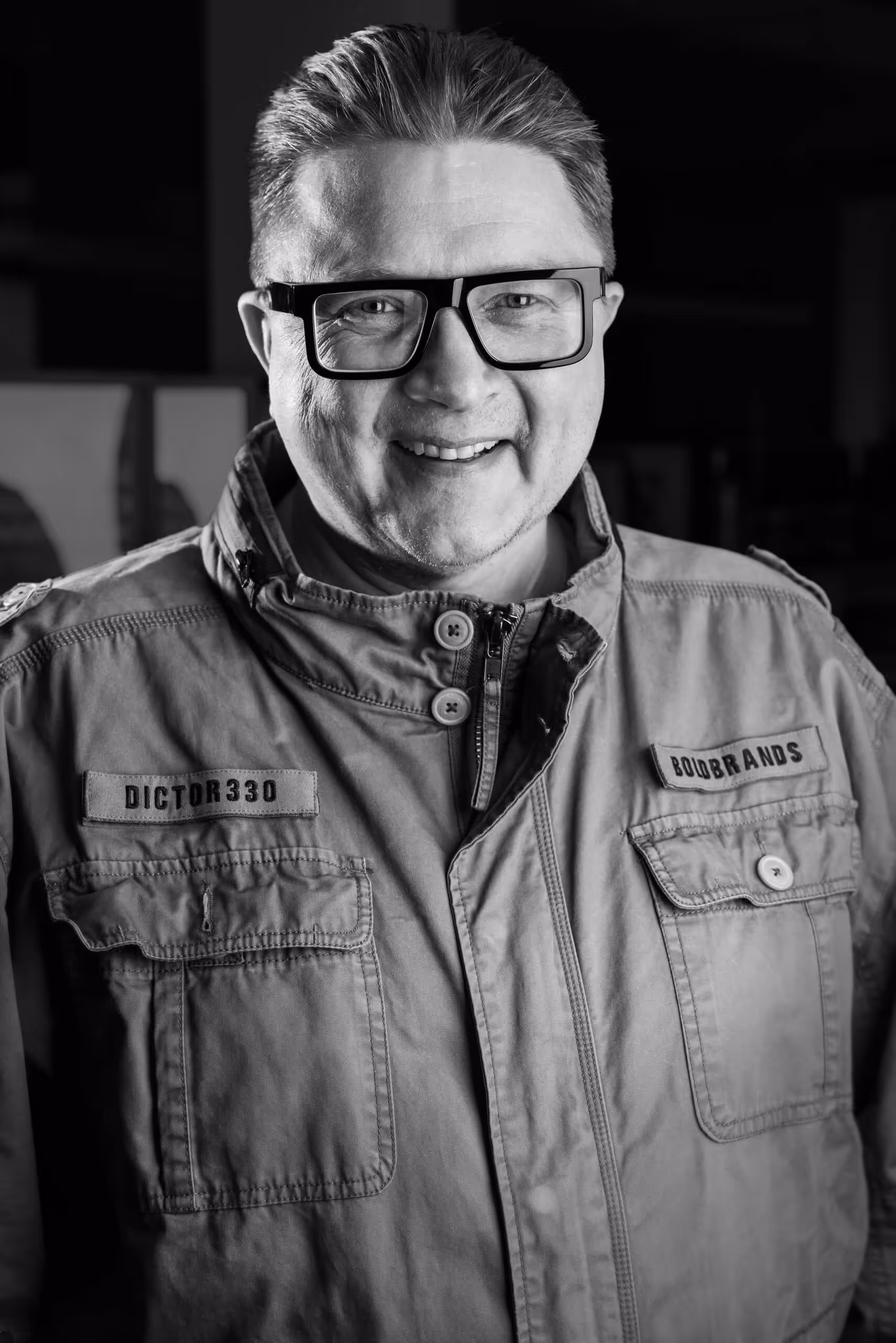 Smiling man with short hair wearing large square glasses and a jacket with patches labeled DICTOR330 and BOIDBRANDS.