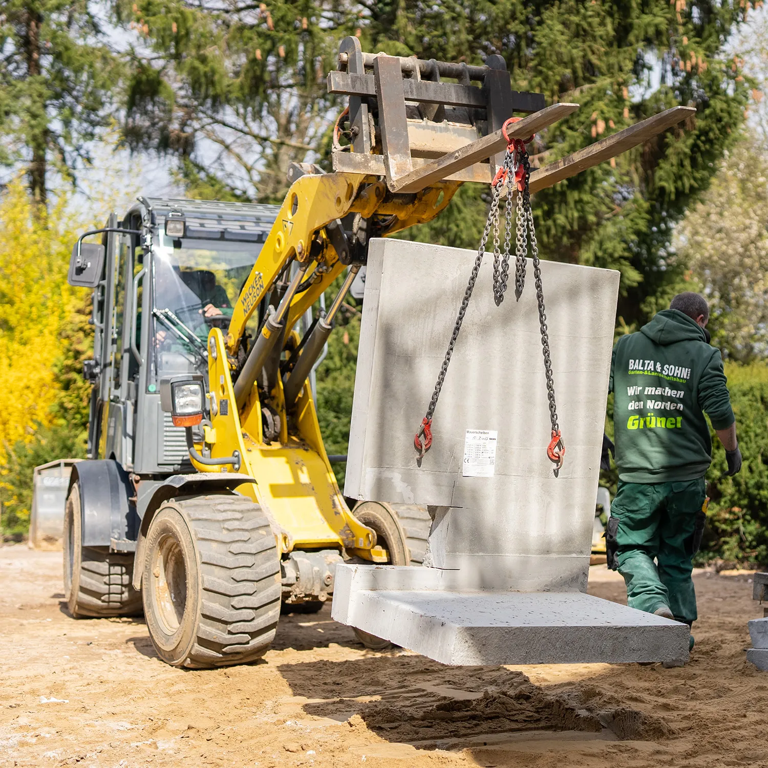 Baustelle mit einem gelben Radlader, der zwei große Betonplatten hebt, und einem Arbeiter in grüner Arbeitskleidung.
