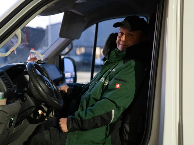 Mann in grüner Jacke und schwarzer Mütze sitzt am Steuer eines Fahrzeugs und lächelt.