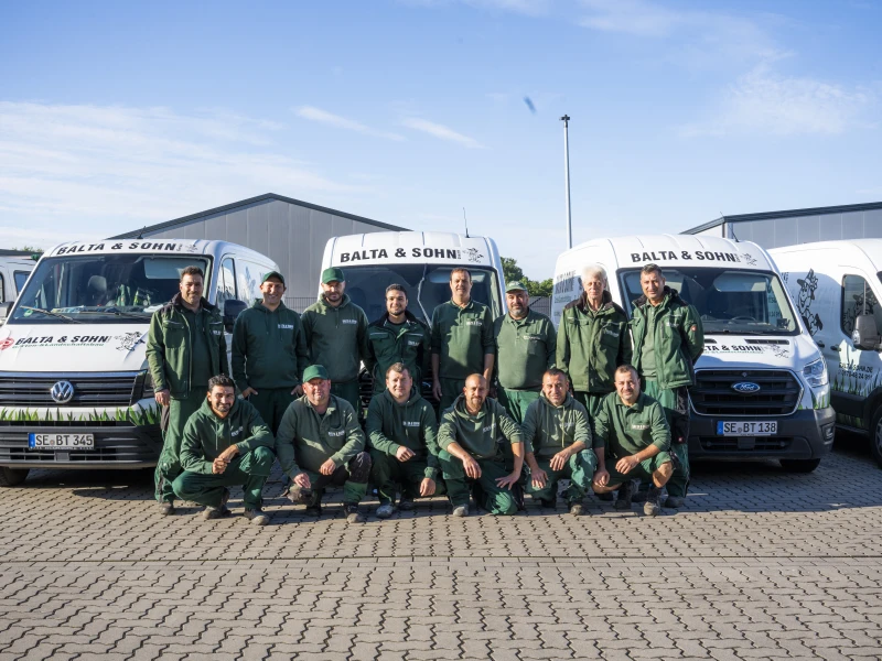 Gruppenfoto von 14 Mitarbeitern in grünen Arbeitskleidung vor drei weißen Firmenfahrzeugen mit der Aufschrift ‚BALTA & SOHN‘.