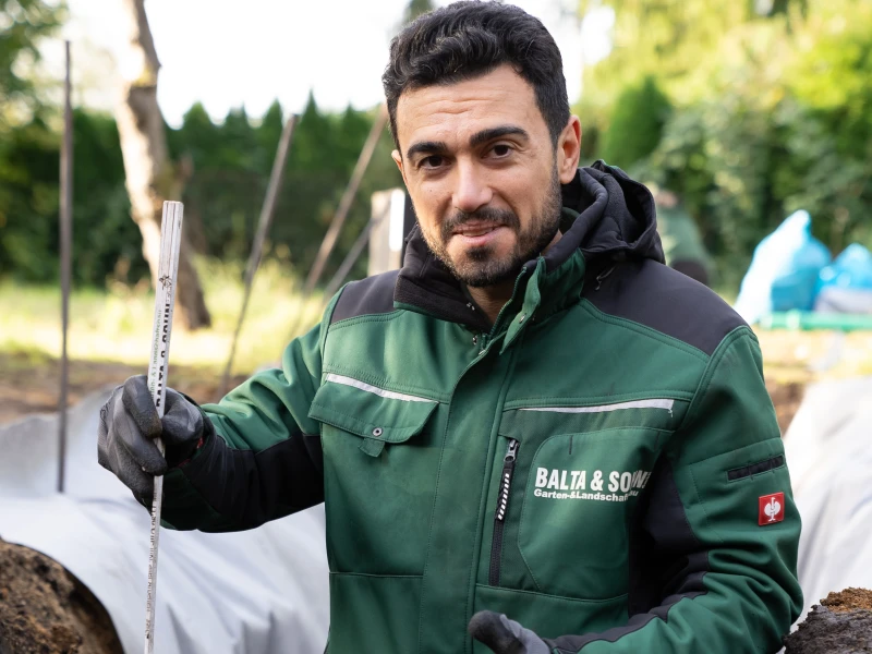 Mann in grüner Arbeitsjacke von Balta & Sohn hält eine Messlatte in einem Garten mit Pflanzen und Baumpfählen.