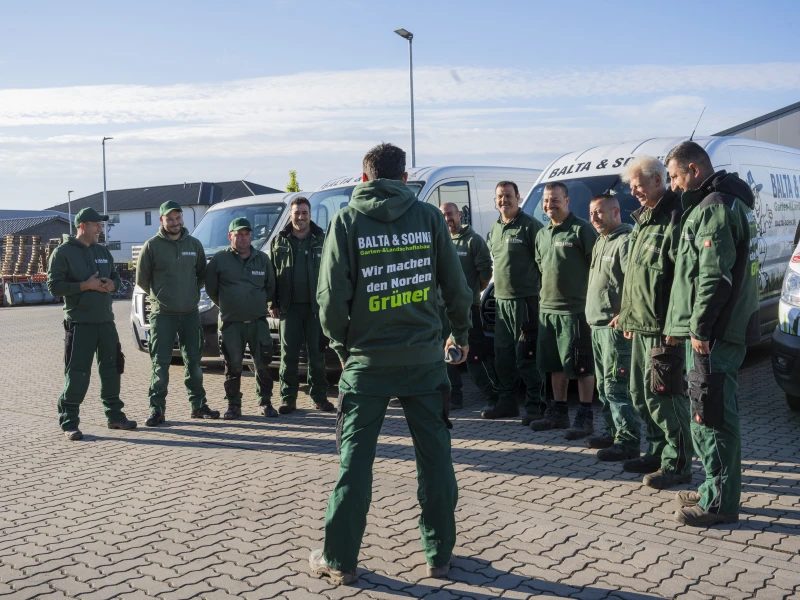 Team von elf Personen in grüner Arbeitskleidung steht im Halbrund vor mehreren Firmenfahrzeugen auf gepflastertem Boden im Freien bei Tageslicht.