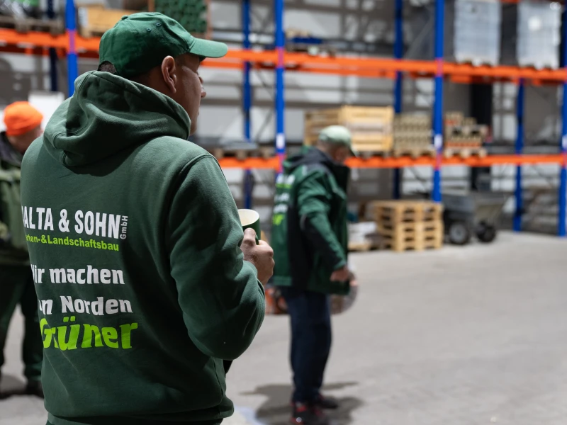 Mann in grünem Kapuzenpullover und grüner Mütze hält einen Becher in einer Lagerhalle mit Regalen und Paletten.