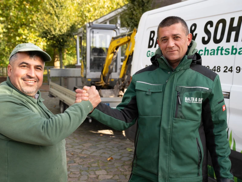 Zwei Männer in grüner Arbeitskleidung geben sich vor einem Firmenfahrzeug mit Bagger in der Nähe die Hand.