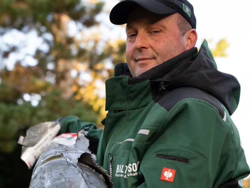 Ein Mann in grüner Arbeitsjacke mit schwarzer Kapuze und schwarzer Mütze, der draußen neben einem beschädigten Rohr steht.