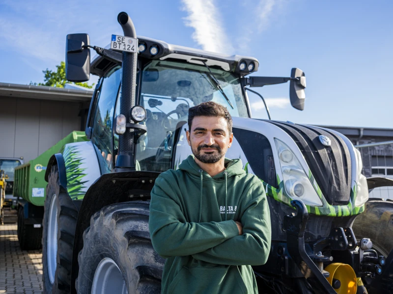 Mann mit grünem Hoodie steht mit verschränkten Armen vor einem großen Traktor unter blauem Himmel.