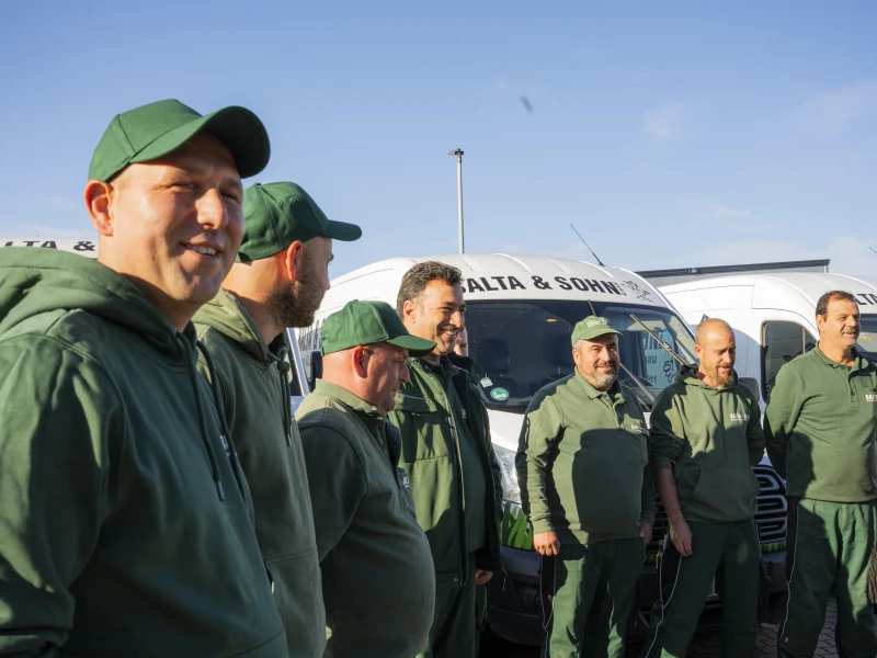 Sieben Männer in grüner Arbeitskleidung stehen vor Firmenfahrzeugen mit der Aufschrift ‚Balta & Sohn‘ unter blauem Himmel.