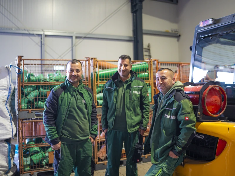 Drei Männer in grüner Arbeitskleidung stehen in einem Lager mit Metallcontainern voller grüner Rohre neben einem gelben Gabelstapler.