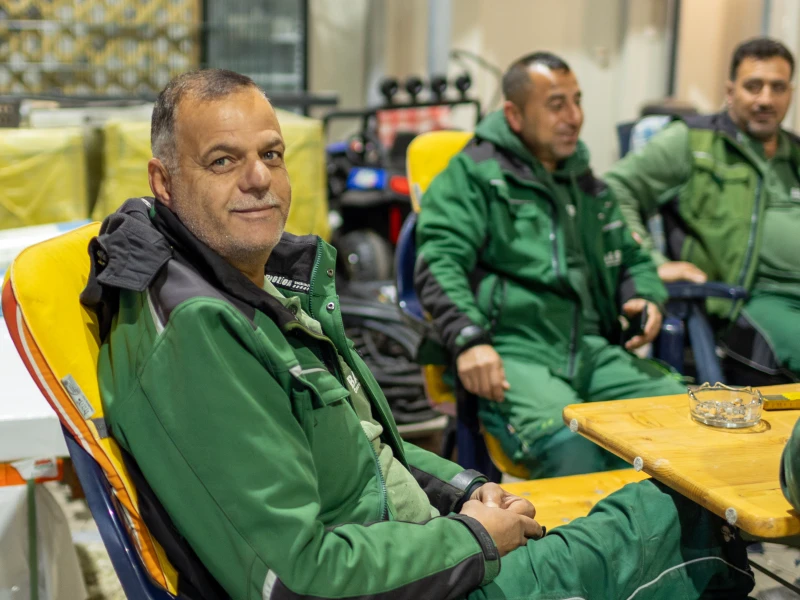 Drei Männer in grüner Arbeitskleidung sitzen entspannt auf Stühlen um einen Holztisch herum.