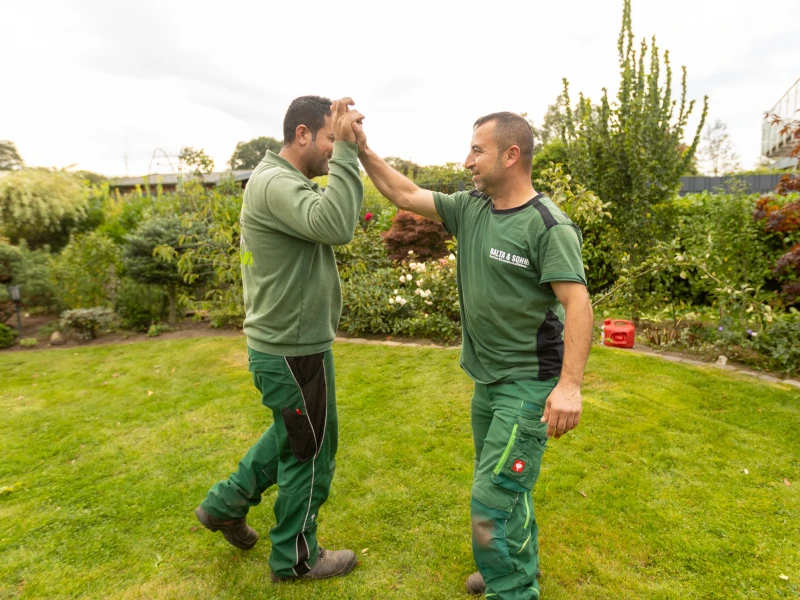Zwei Gärtner in grüner Arbeitskleidung geben sich im Garten einen High-Five.