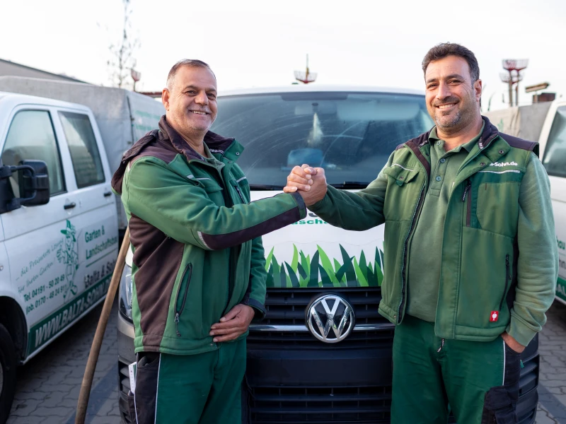 Zwei Männer in grüner Arbeitskleidung geben sich vor einem Firmenfahrzeug mit Volkswagen-Logo die Hand.