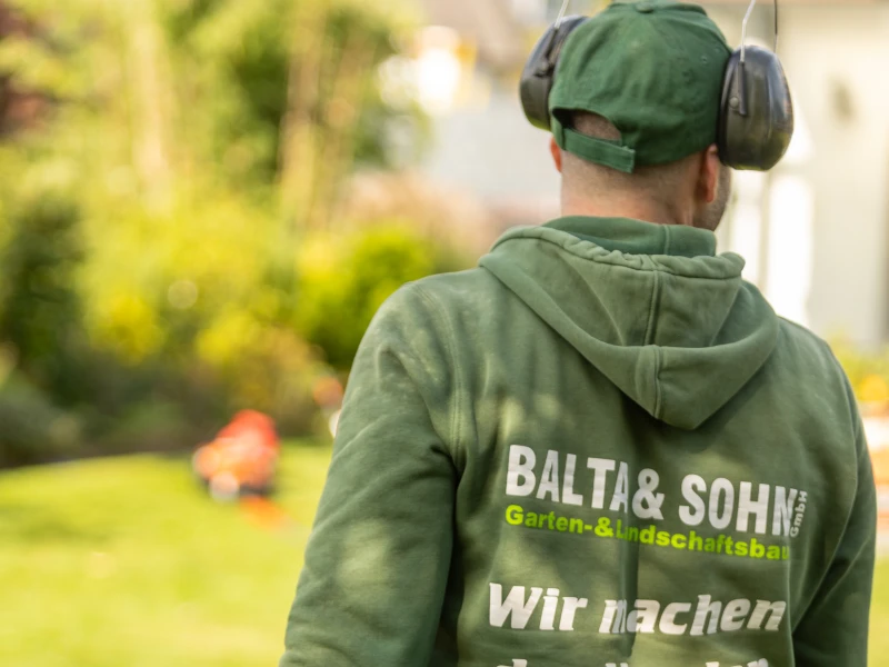 Mann mit grünem Kapuzenpullover und grüner Kappe mit Gehörschutz im Garten, auf dessen Rücken ‚BALTA & SOHN Garten- & Landschaftsbau GmbH‘ steht.