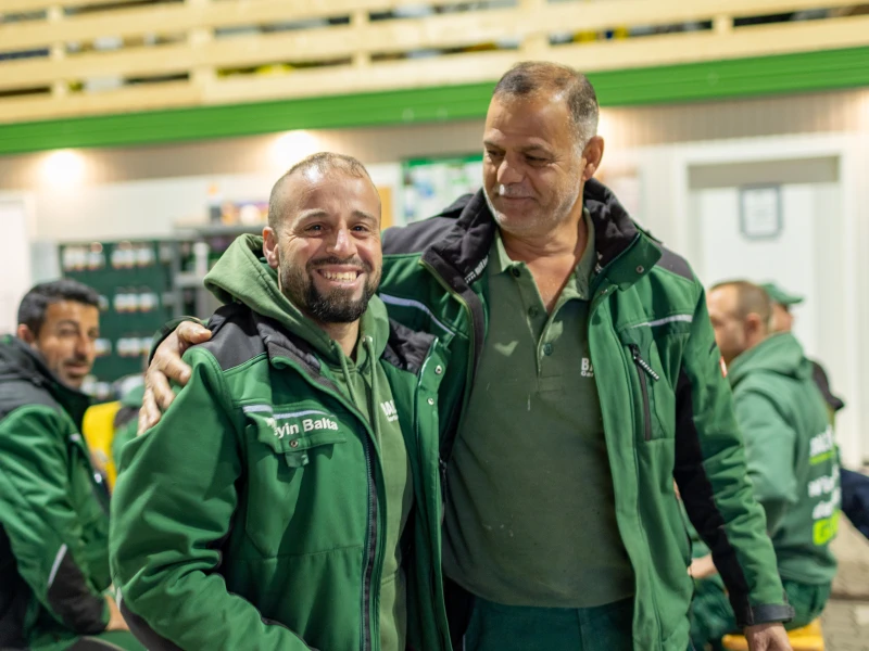 Zwei Männer in grünen Arbeitsjacken umarmen sich und lächeln in einer Werkstatt oder Lagerhalle.