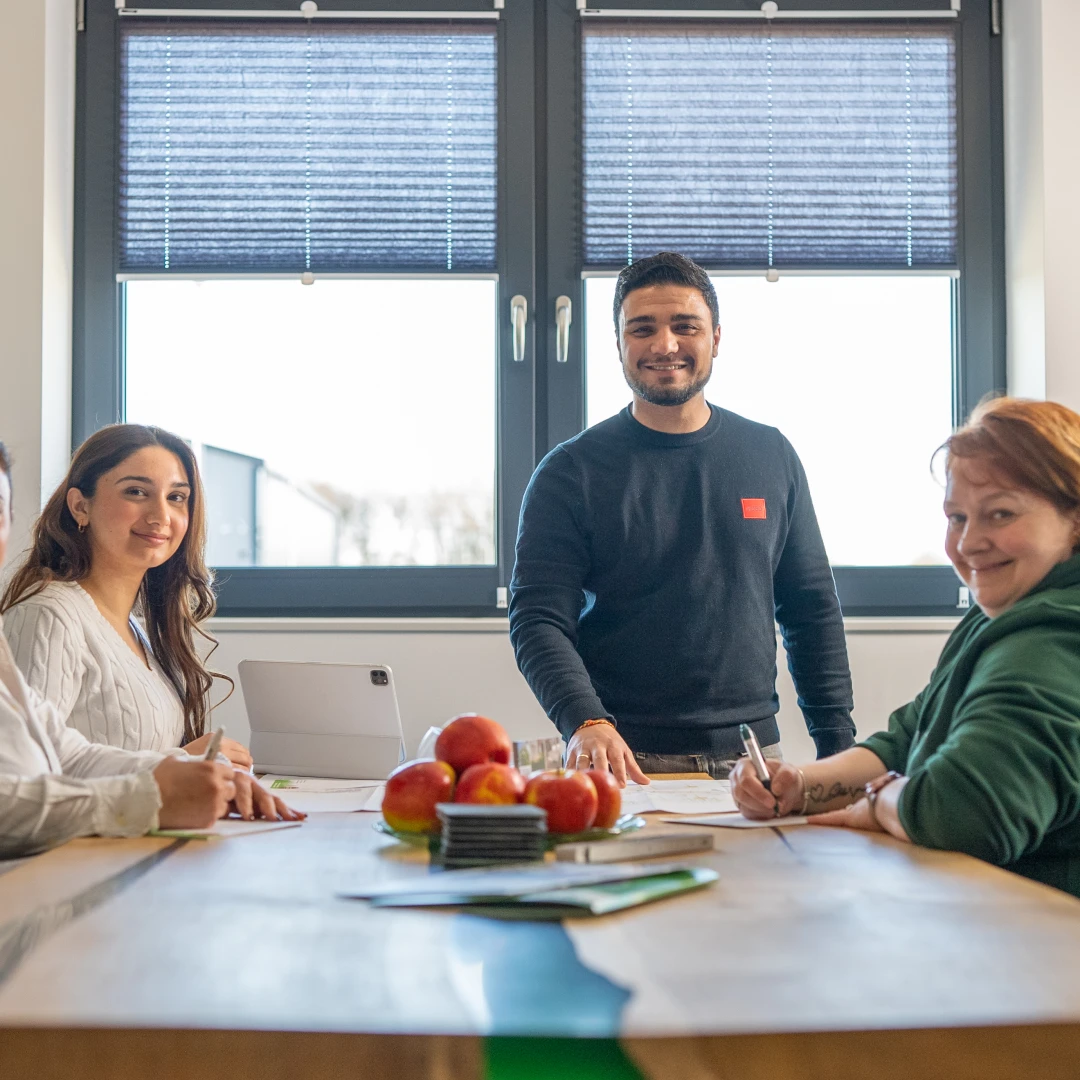 Drei Personen sitzen an einem Tisch mit Äpfeln und Unterlagen vor einem Fenster, eine Person steht und lächelt.