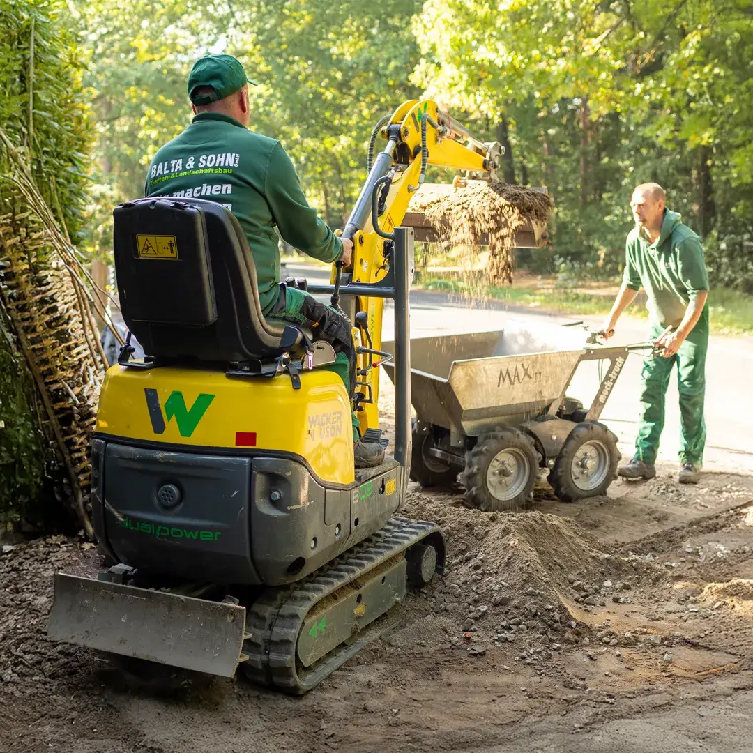 Zwei Männer bei Erdarbeiten, einer bedient einen gelben Minibagger und kippt Erde in einen Kipplader, umgeben von Bäumen.