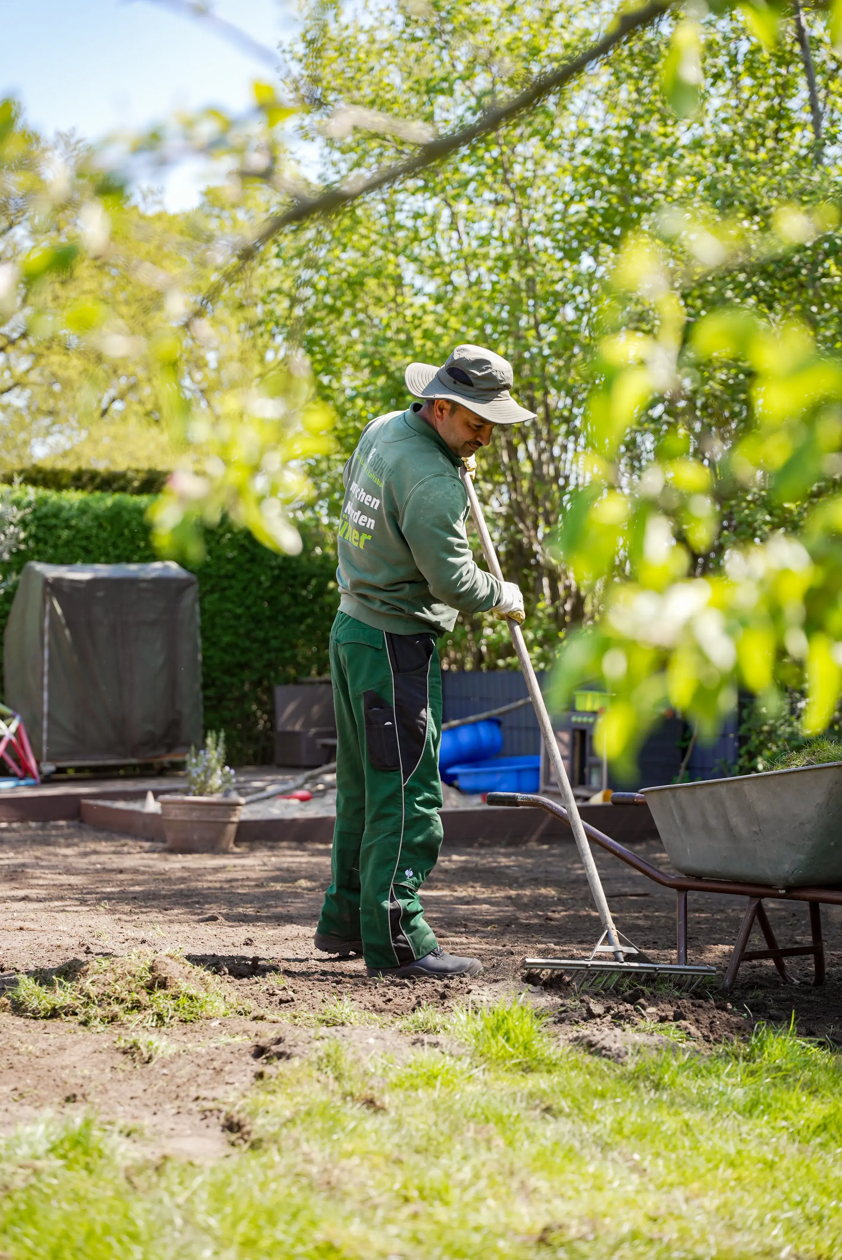 Gärtner in grüner Kleidung und Hut harkt Erde in einem Garten bei sonnigem Wetter.