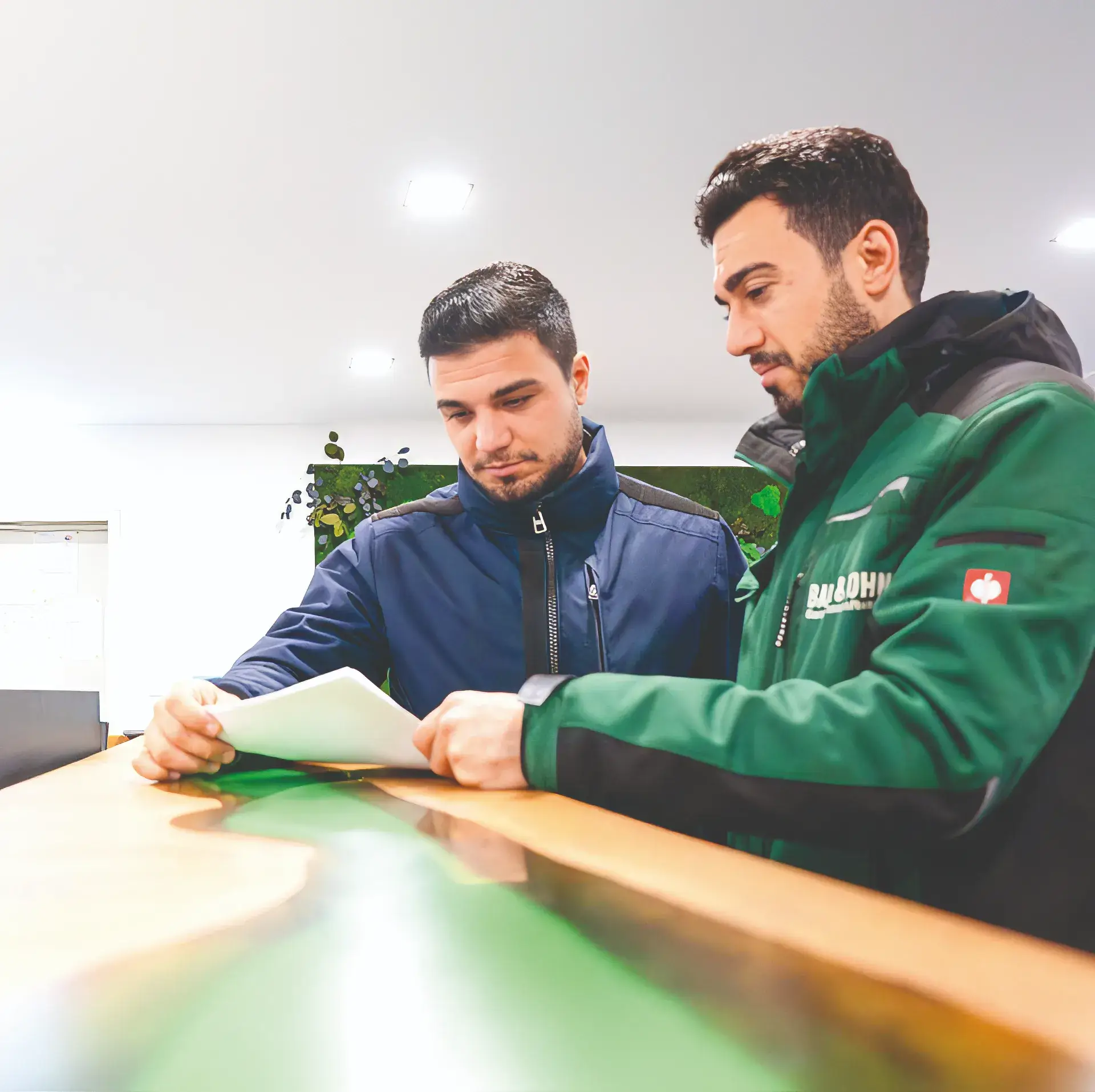 Zwei Männer in Outdoorjacken diskutieren über ein Blatt Papier an einem Holz- und grünen Tisch.