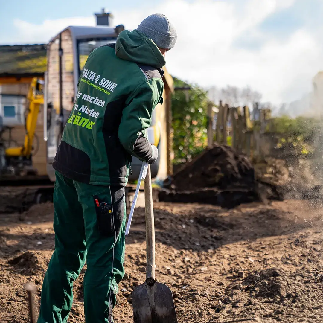 Gärtner in grüner Arbeitskleidung misst mit Maßband eine Bodenfläche auf einer Baustelle mit Schaufel.