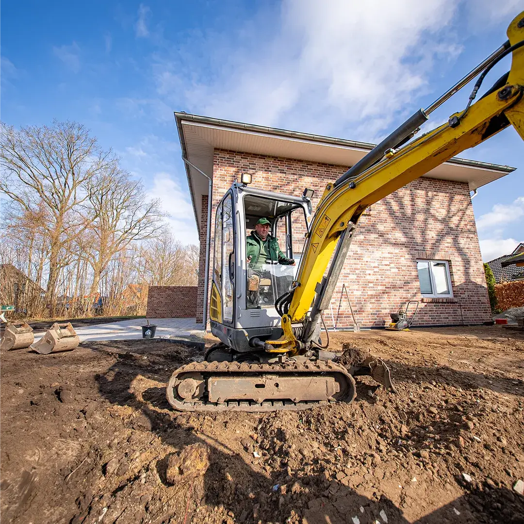 Baggerfahrer arbeitet mit kleinem gelben Bagger vor einem Backsteinhaus an einer Baustelle unter blauem Himmel.