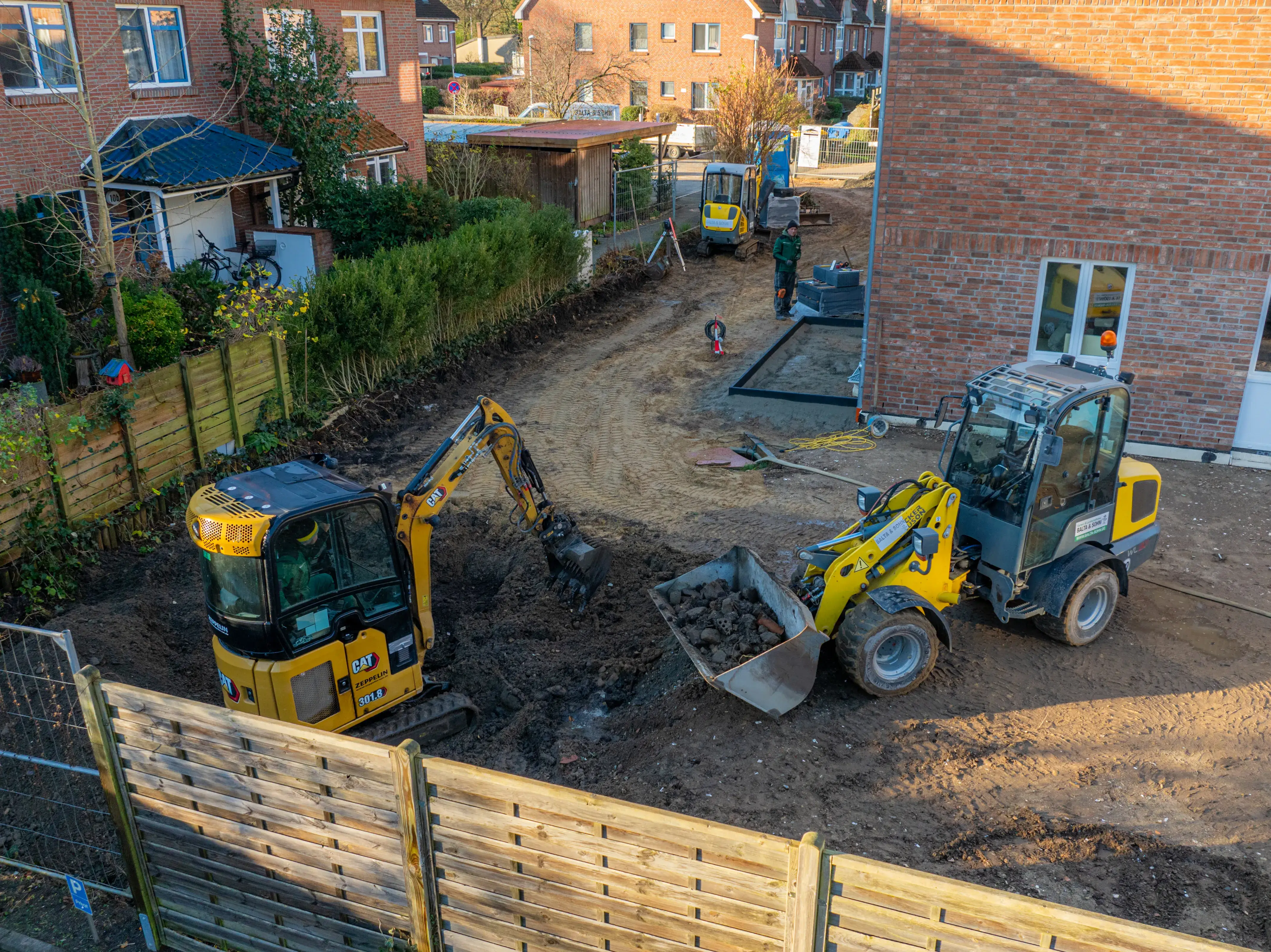 Baustelle mit zwei gelben Baggern und einem Arbeiter vor einem Wohnhaus mit rotbraunen Ziegeln.