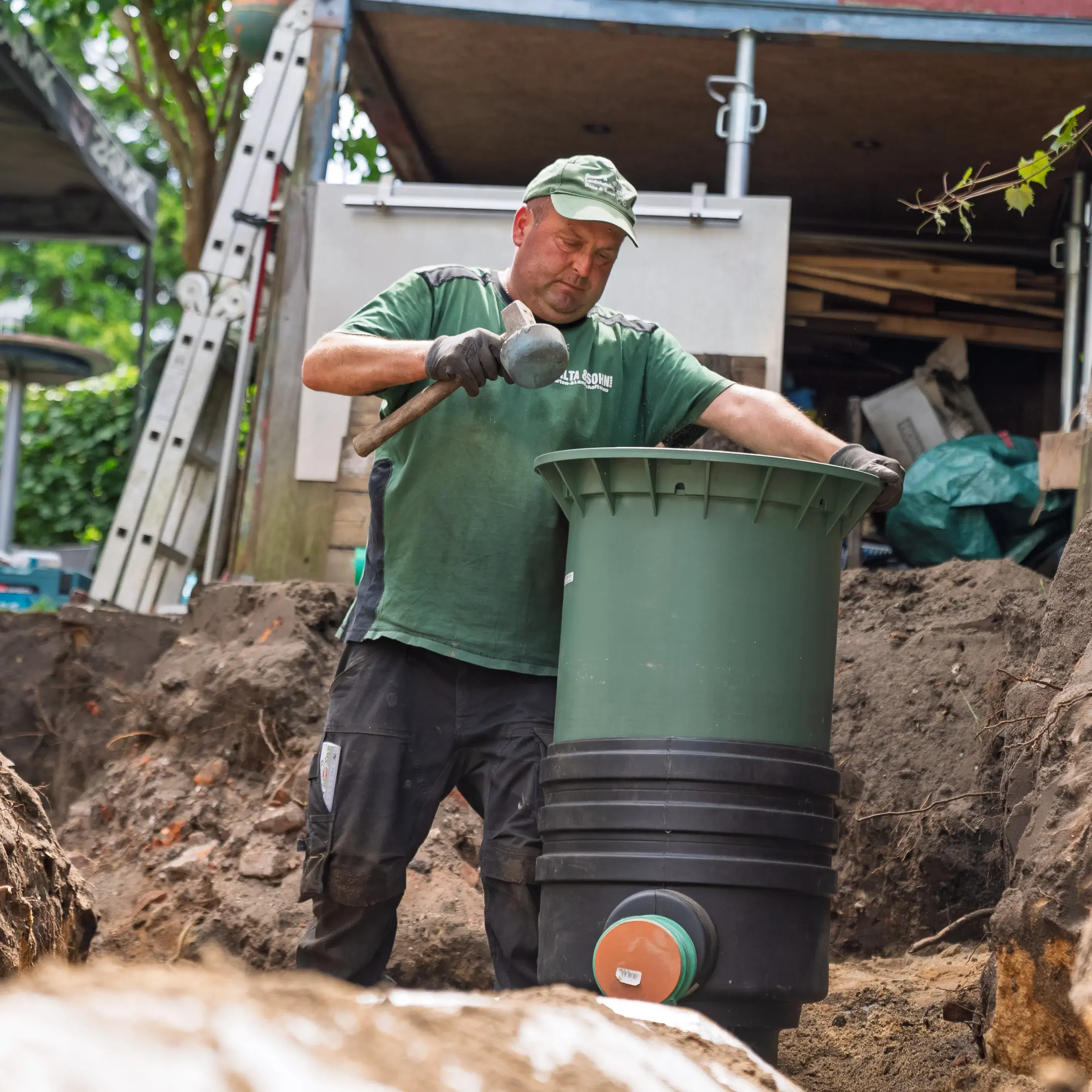 Arbeiter in grüner Kleidung installiert eine große grüne Kunststoffanlage im ausgegrabenen Erdreich.