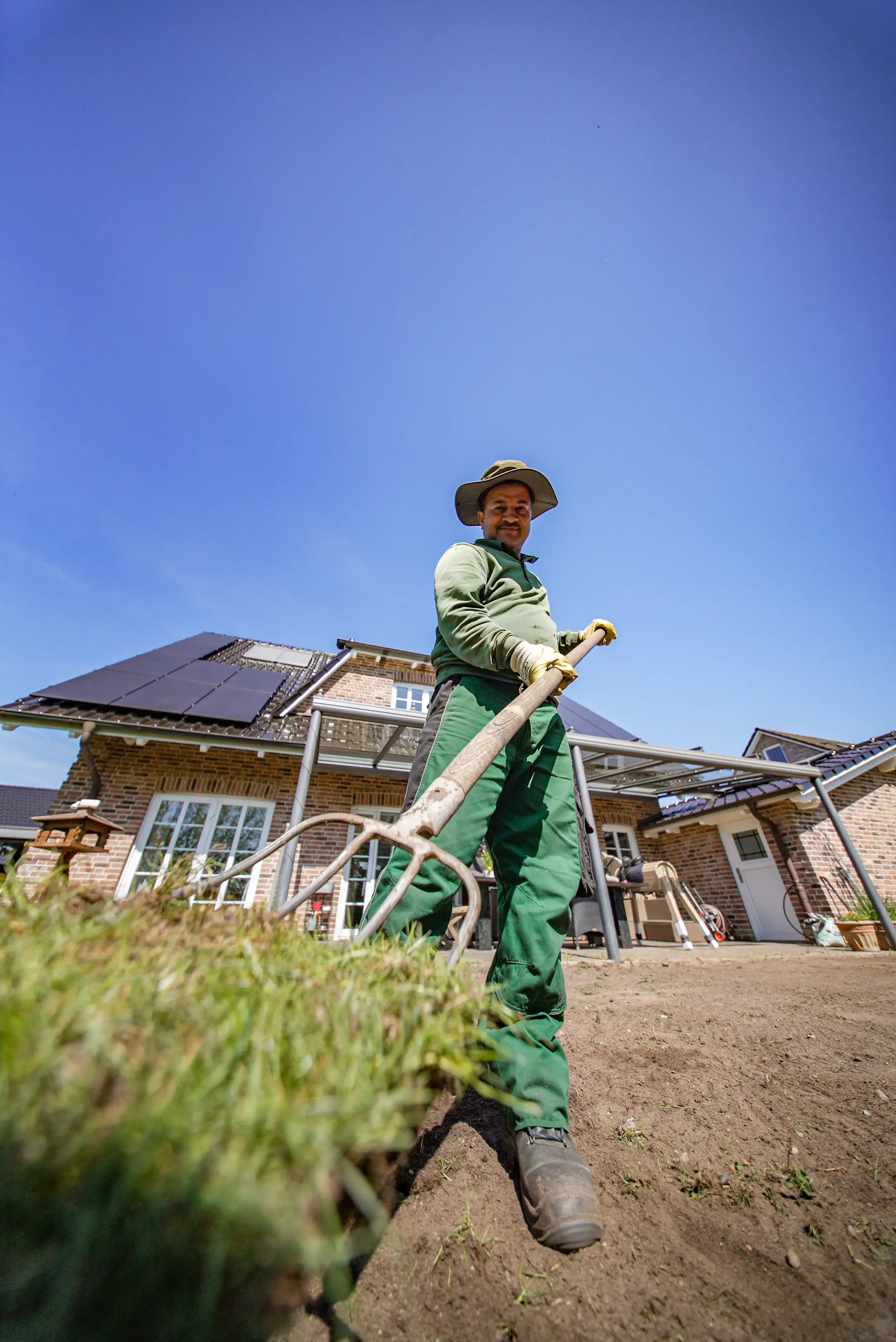 Gärtner mit grünem Arbeitsanzug und Hut steht auf einem frisch bearbeiteten Gartenboden vor einem Haus mit Solarpanelen und hält eine Hacke.